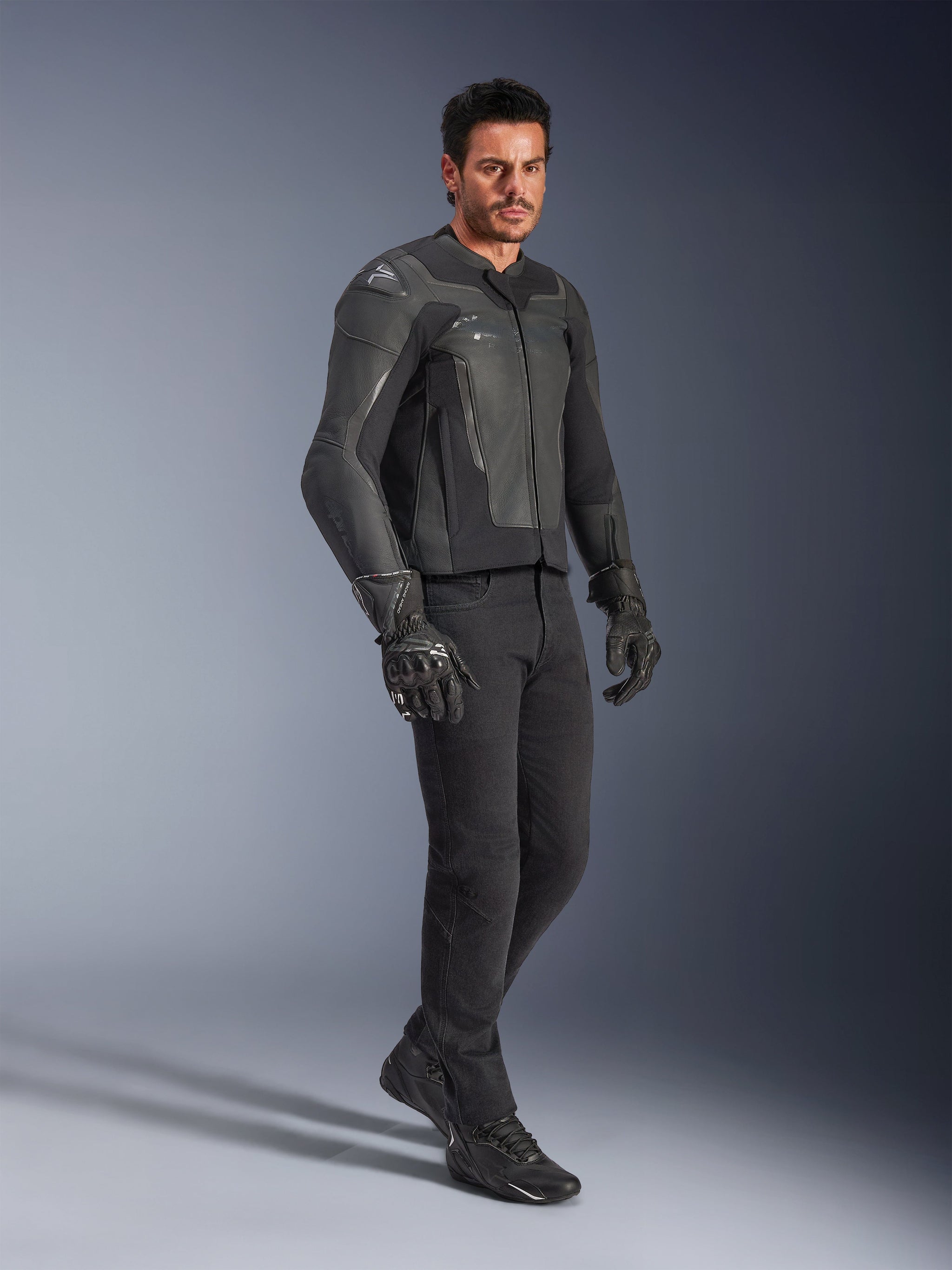 Man wearing Alpinestars GP Force V2 Leather Jacket in Black, paired with black pants and motorcycle gloves, full-body studio shot, sleek bovine leather construction with integrated shoulder and elbow protection, professional sport motorcycle road gear