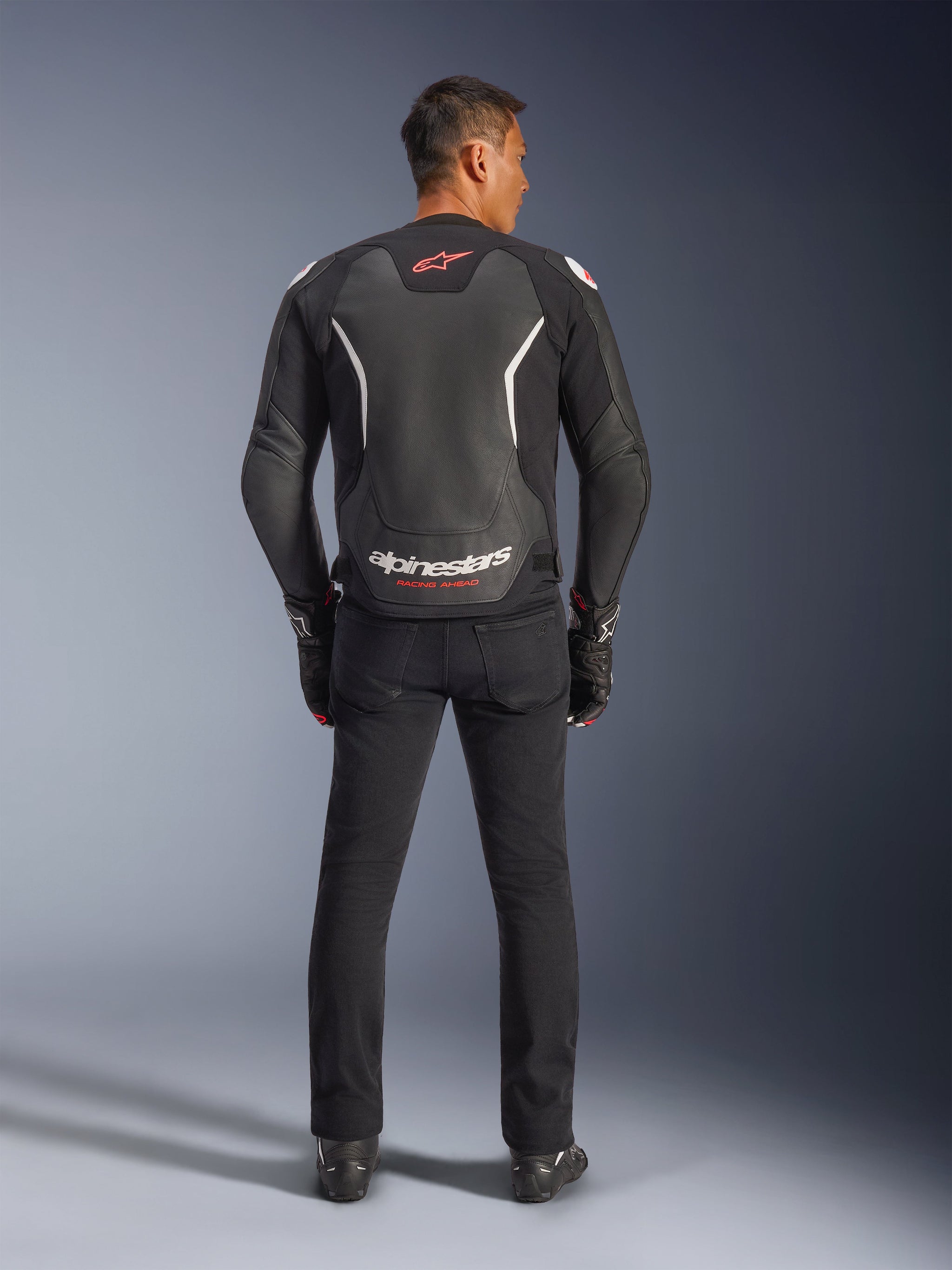 Alpinestars GP Force V2 Leather Jacket, Black and White, rear view, worn by a rider with black pants and gloves, featuring large white branding on the lower back and red accents, premium bovine leather construction for motorcycle road gear