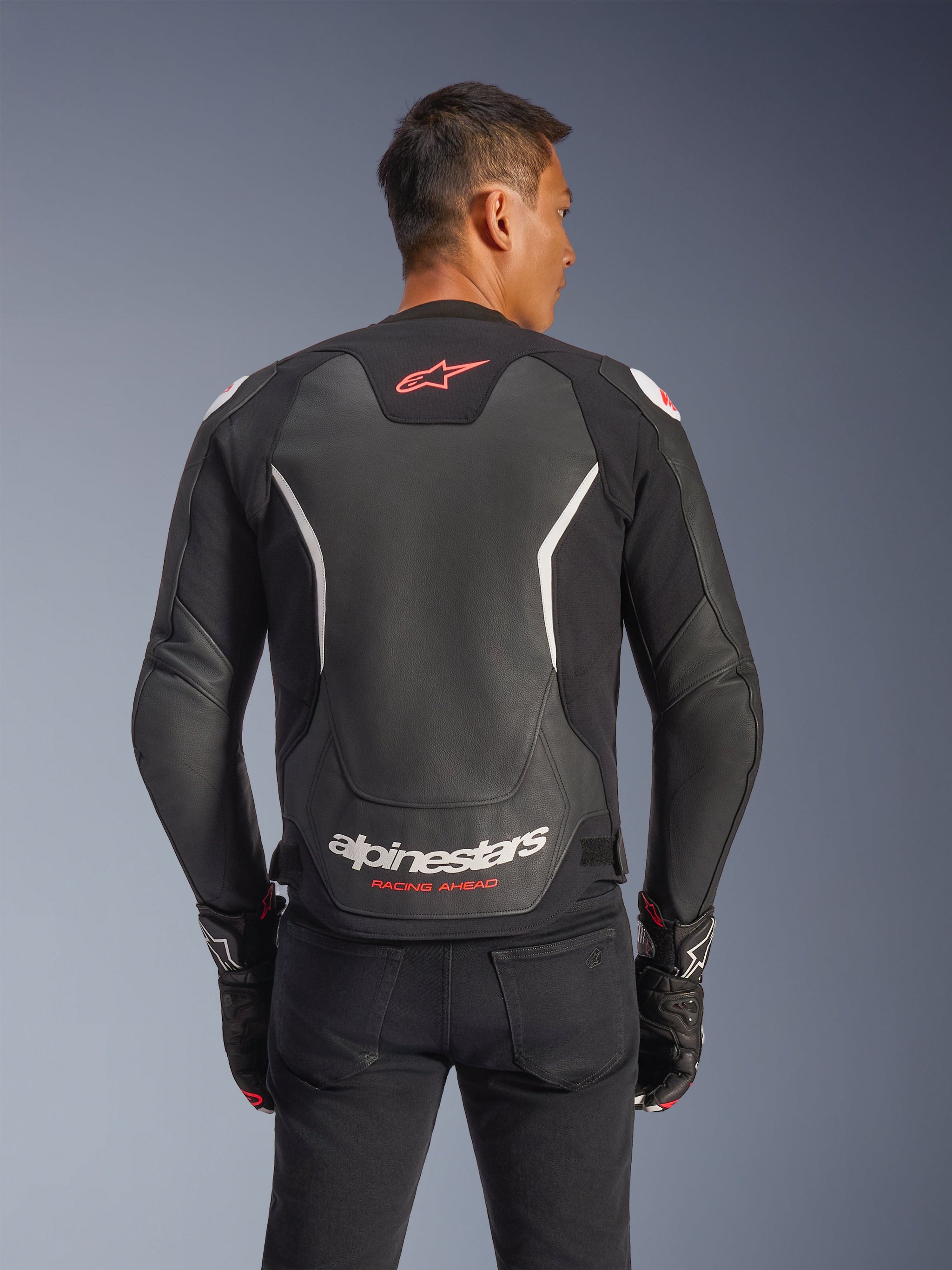 Alpinestars GP Force V2 Leather Jacket, Rear view, Black and White, featuring red star logo on the upper back and white Alpinestars branding with 'Racing Ahead' text, worn with black pants and matching gloves, sport-fit leather motorcycle jacket