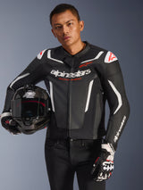 Alpinestars GP Force V2 Leather Jacket, Black and White with red accents, worn by a model holding a carbon fiber motorcycle helmet and wearing racing gloves, featuring white contrast panels and Alpinestars branding.