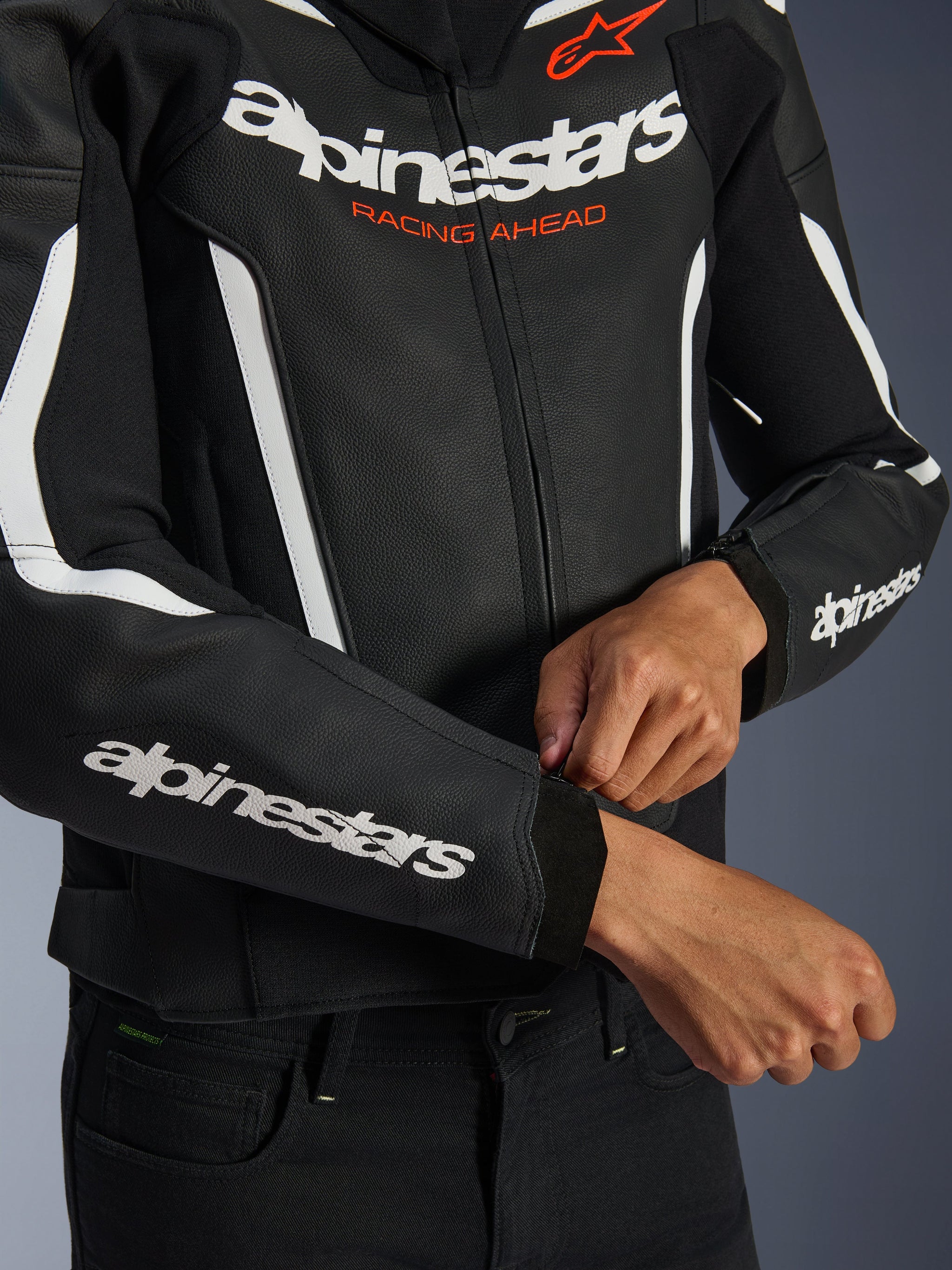 Alpinestars GP Force V2 Leather Jacket, Black and White, close-up of rider adjusting the cuff zipper, white branding on chest and sleeves with orange 'Racing Ahead' text, premium bovine leather for sport motorcycle riding