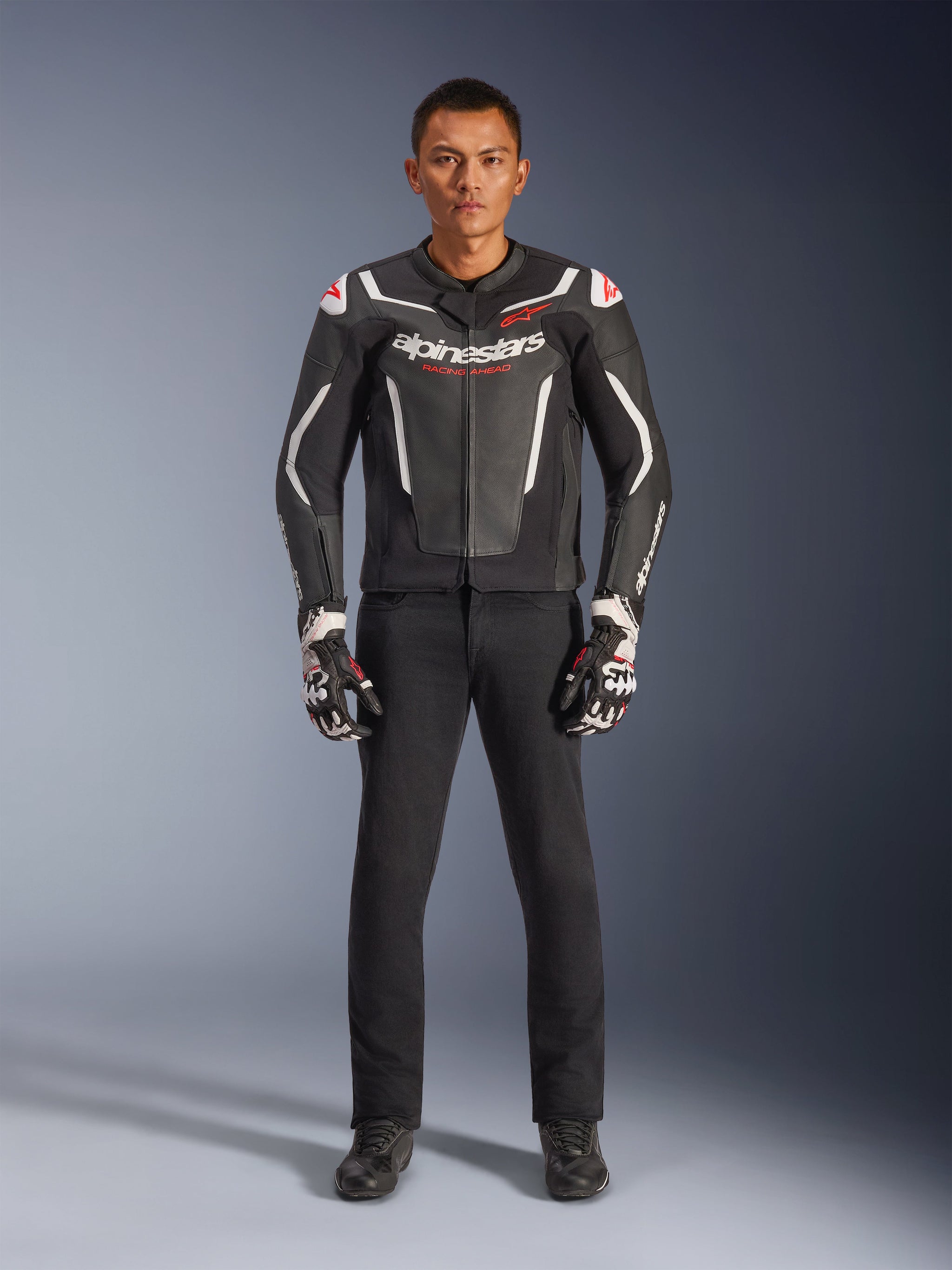 Male model wearing Alpinestars GP Force V2 Leather Jacket in Black and White, paired with black motorcycle pants and black and white gloves, featuring bold Alpinestars branding and red shoulder accents, professional sport motorcycle riding gear