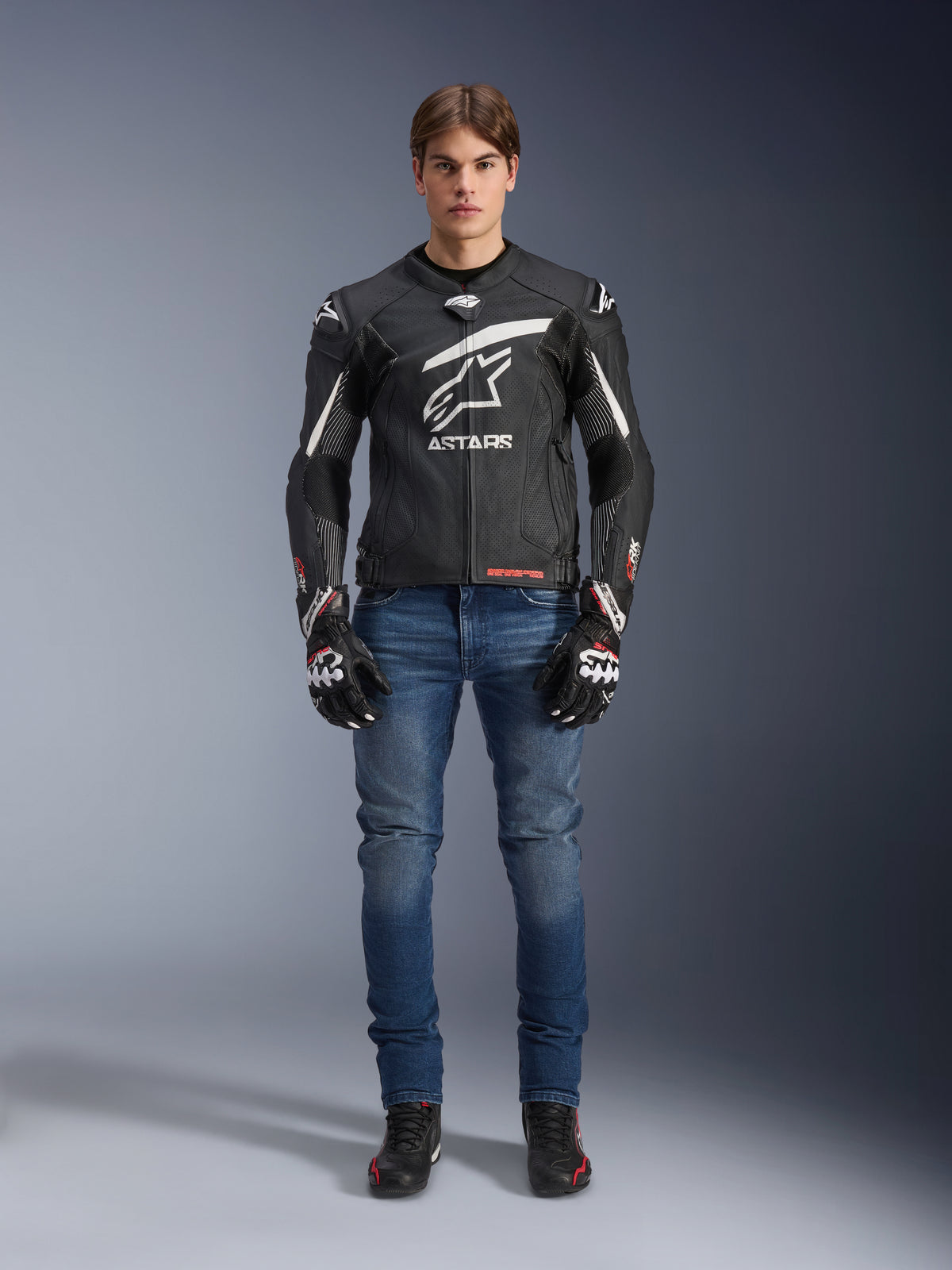 Male model wearing Alpinestars GP Plus R V4 Rideknit Leather Jacket in Black and White, styled with blue riding jeans, black motorcycle gloves, and black riding shoes, featuring a large white Astars logo on the chest, perforated leather panels, and white racing stripes on the sleeves, full-body standing view
