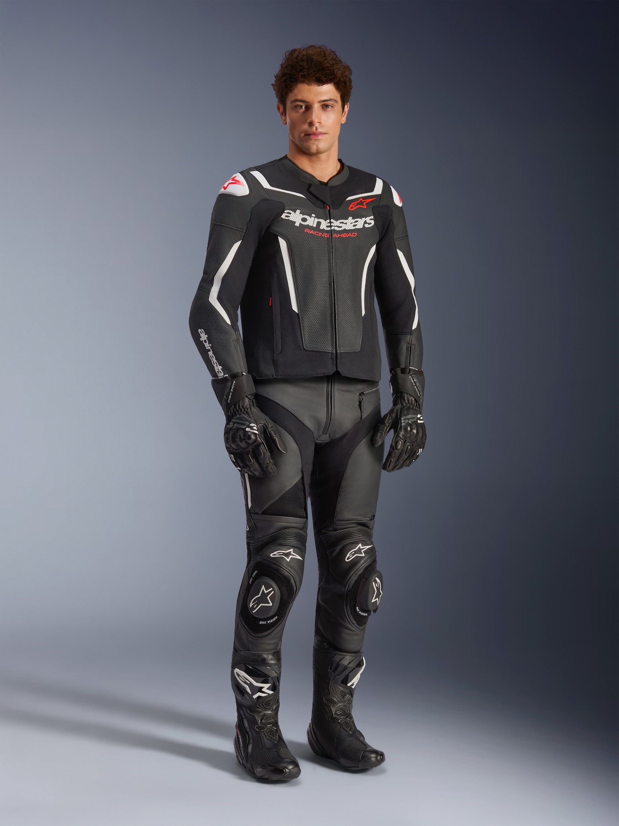 Alpinestars GP Force V2 Air Leather Jacket in Black and White, worn by a male rider with matching black racing pants, gloves, and boots, featuring perforated leather panels, white accents, and red Alpinestars logo on the chest, sport motorcycle riding gear