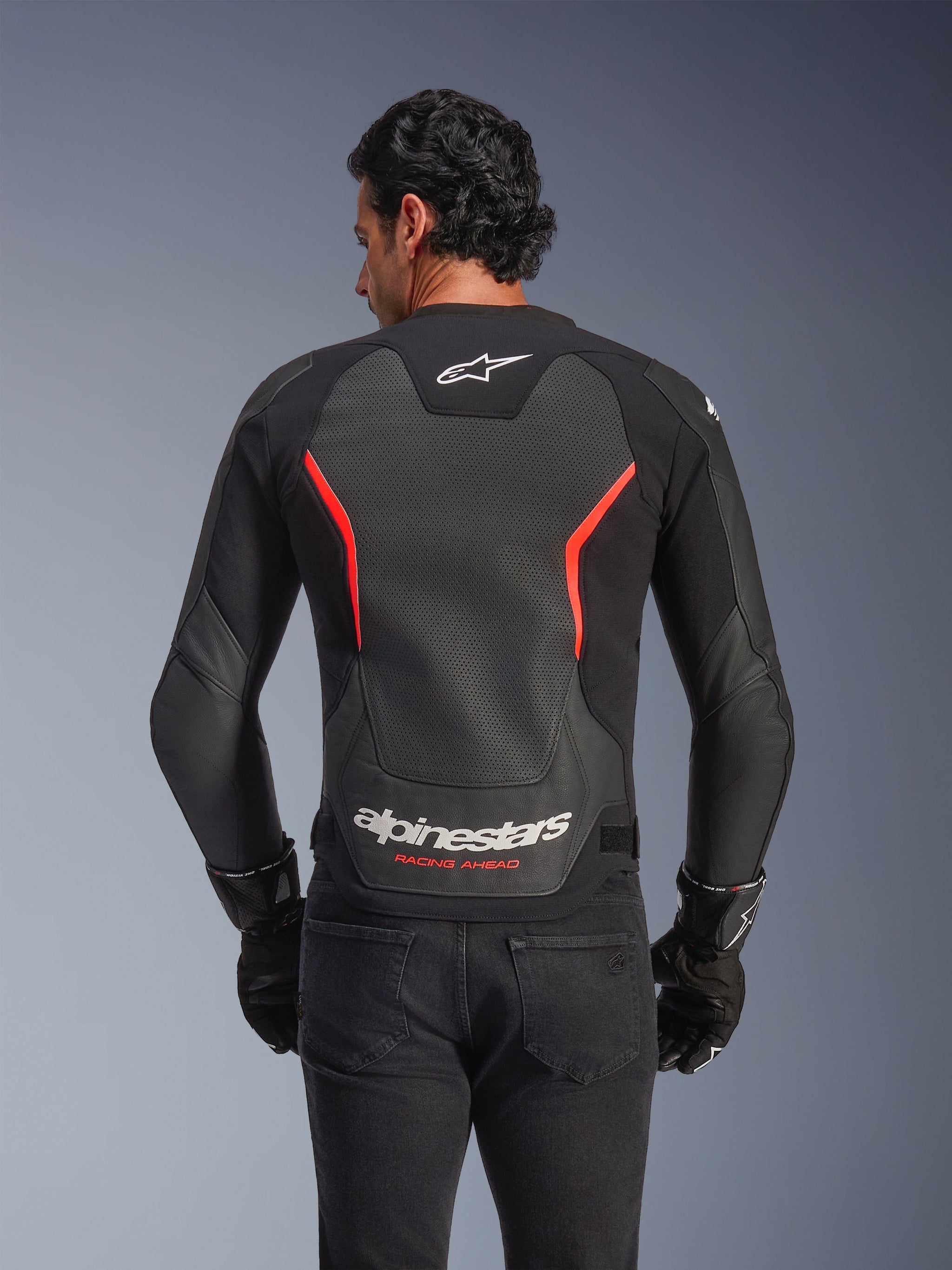Alpinestars GP Force V2 Air Leather Jacket, back view, Black and Red Fluo, featuring a large perforated leather back panel for ventilation, 'Alpinestars Racing Ahead' branding on the lower back, worn by a rider with black gloves, performance sport motorcycle gear