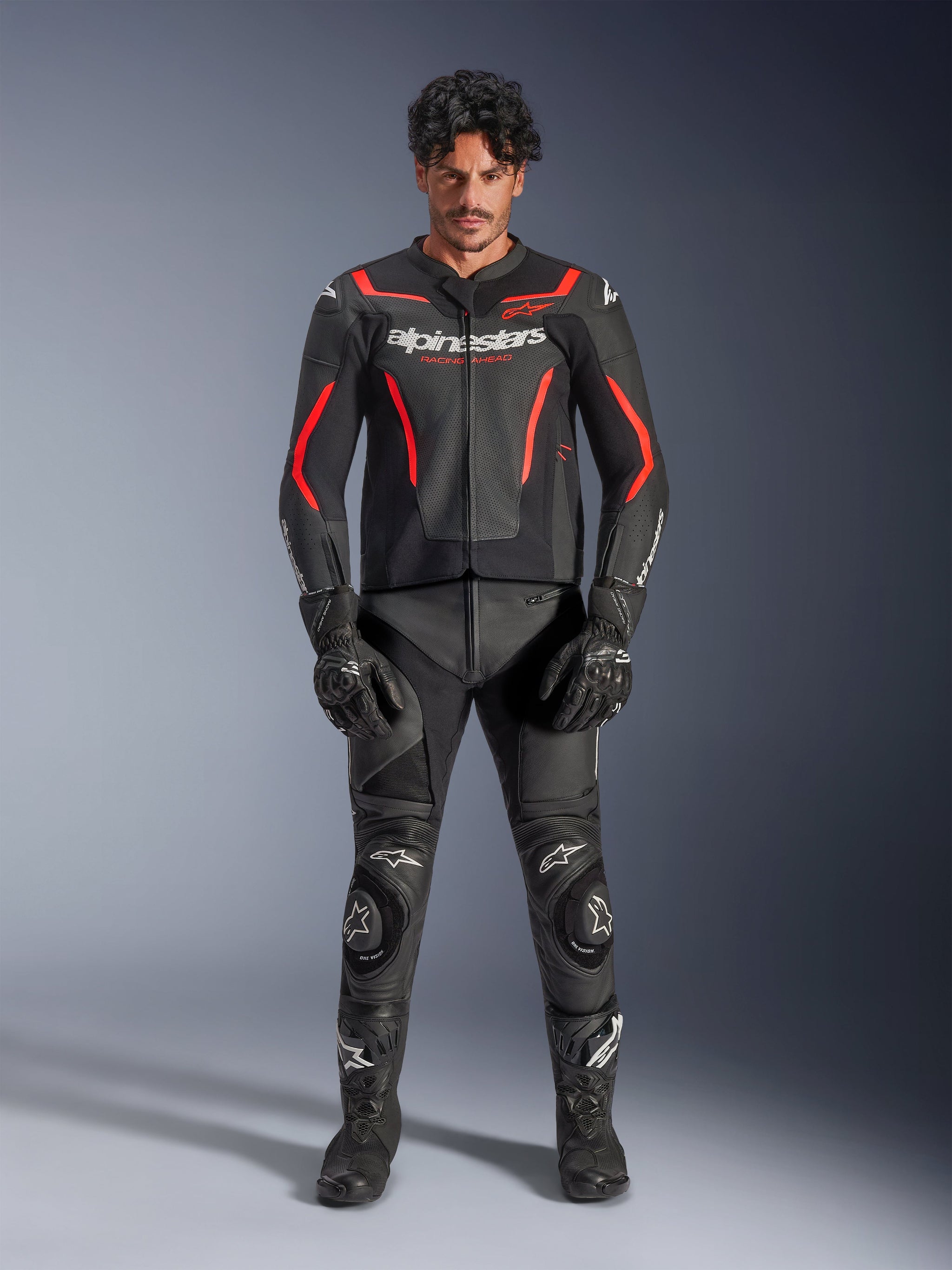 Male model wearing Alpinestars GP Force V2 Air Leather Jacket in Black and Red Fluo, featuring perforated chest panels and white branding, paired with black motorcycle leather pants, black gloves, and black boots for a complete racing look.