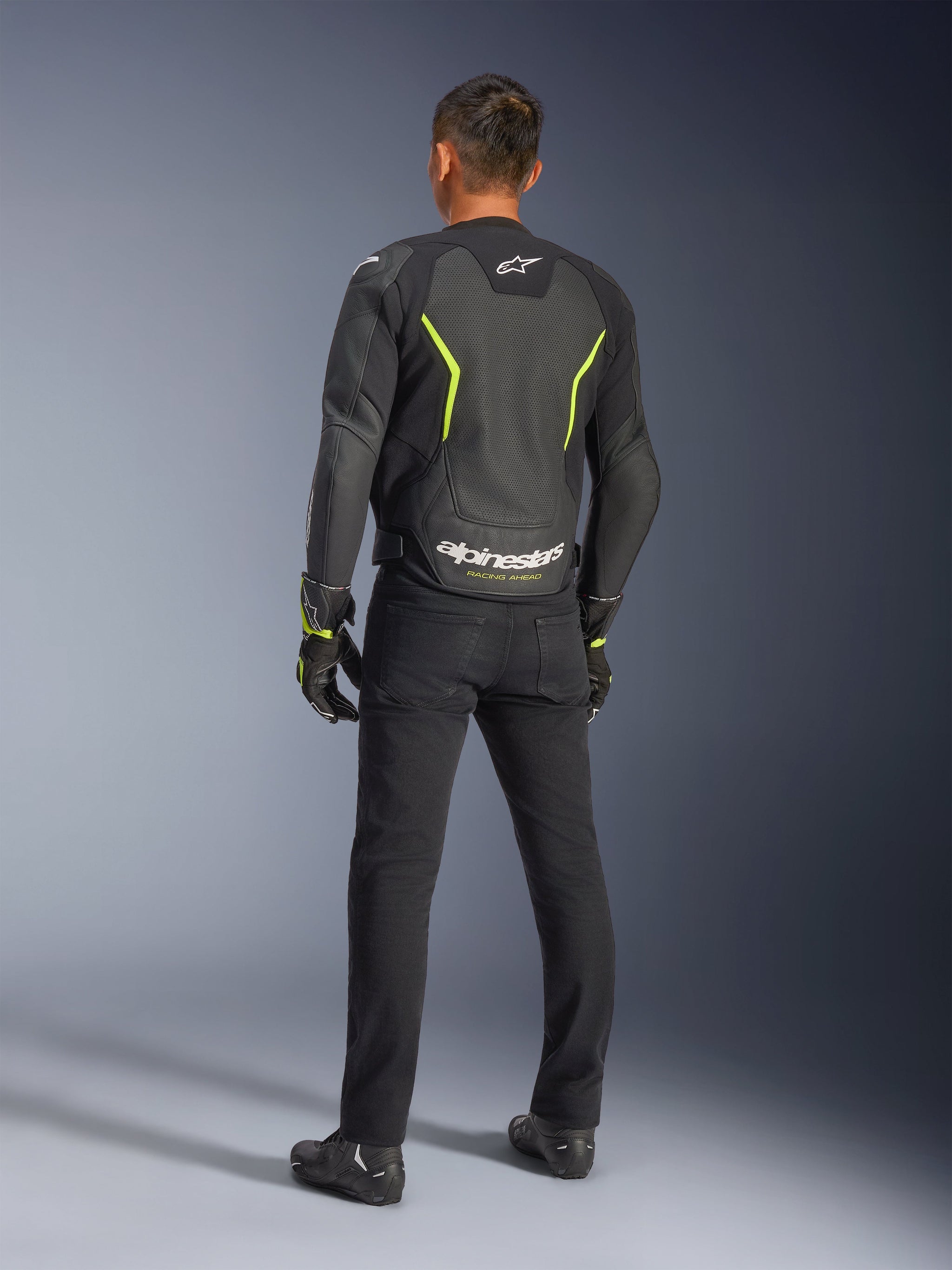Alpinestars GP Force V2 Air Leather Jacket, Black and Yellow Fluo, back view of a rider wearing the perforated leather jacket, featuring angular yellow accents and bold white Alpinestars logo with 'RACING AHEAD' on the lower back, paired with black riding pants and boots.