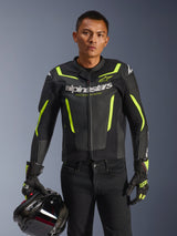 Male rider wearing Alpinestars GP Force V2 Air Leather Jacket in Black and Yellow Fluo, holding a black motorcycle helmet, features perforated leather with bold white Alpinestars branding and neon yellow accents, paired with matching racing gloves and black pants
