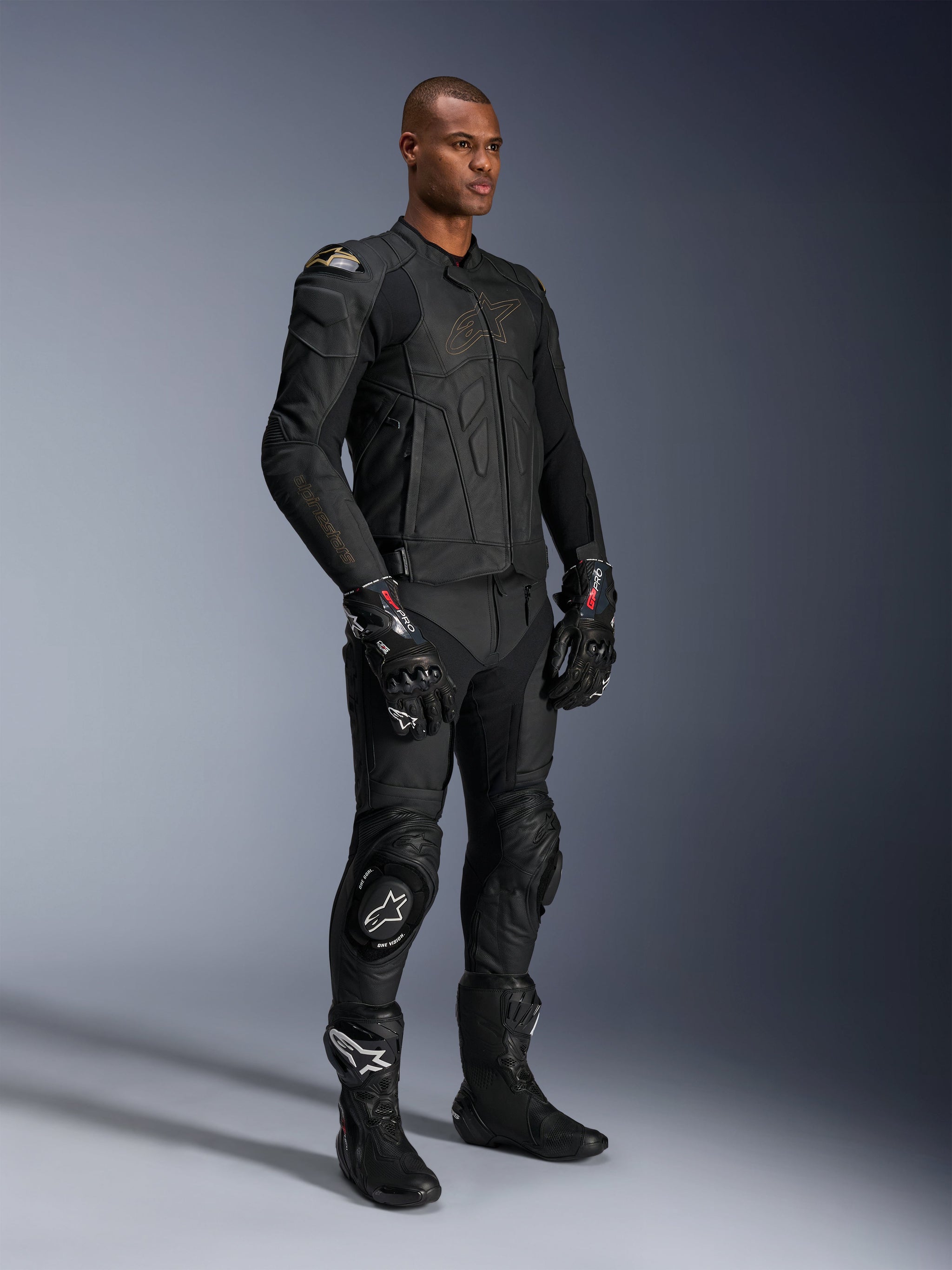 Male model wearing Alpinestars Missile V3 Leather Pants and matching black leather jacket with gold accents, black GP Pro racing gloves, and black racing boots, complete motorcycle racing gear for road use, studio shot.