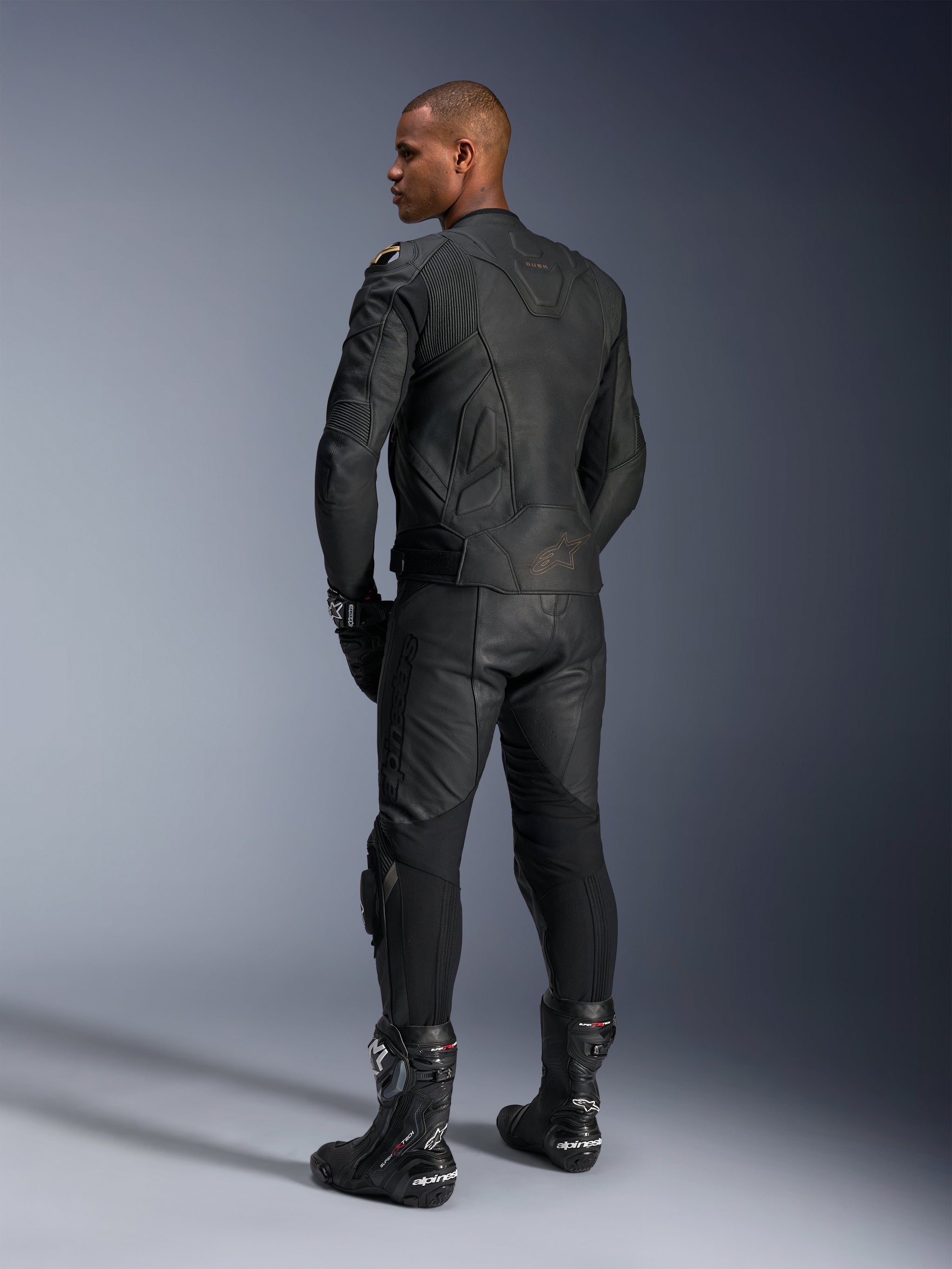 Alpinestars Missile V3 Leather Pants and Dusk Leather Jacket, Black with gold accents, rear view of rider, featuring embossed branding and accordion stretch panels, worn with black Supertech R boots and gloves, professional motorsport apparel.