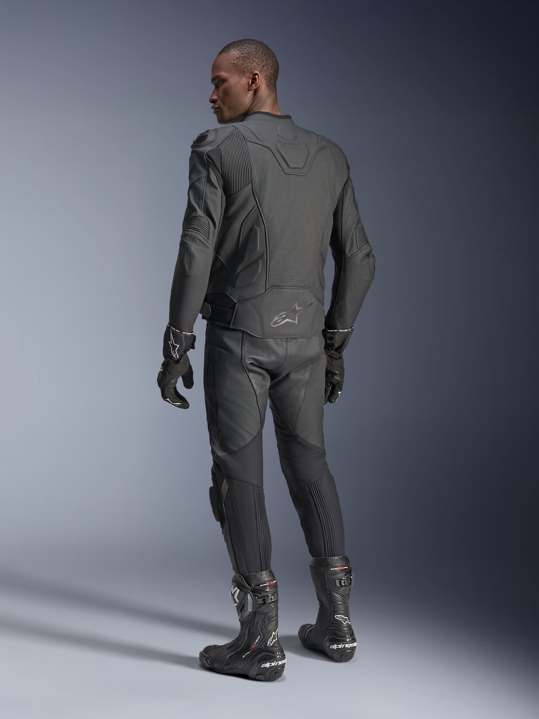 Male rider from behind wearing the Alpinestars Dusk Airflow Leather Jacket in Black Black, paired with matching leather pants, gloves, and boots, highlighting the extensive back perforations and performance sport fit.