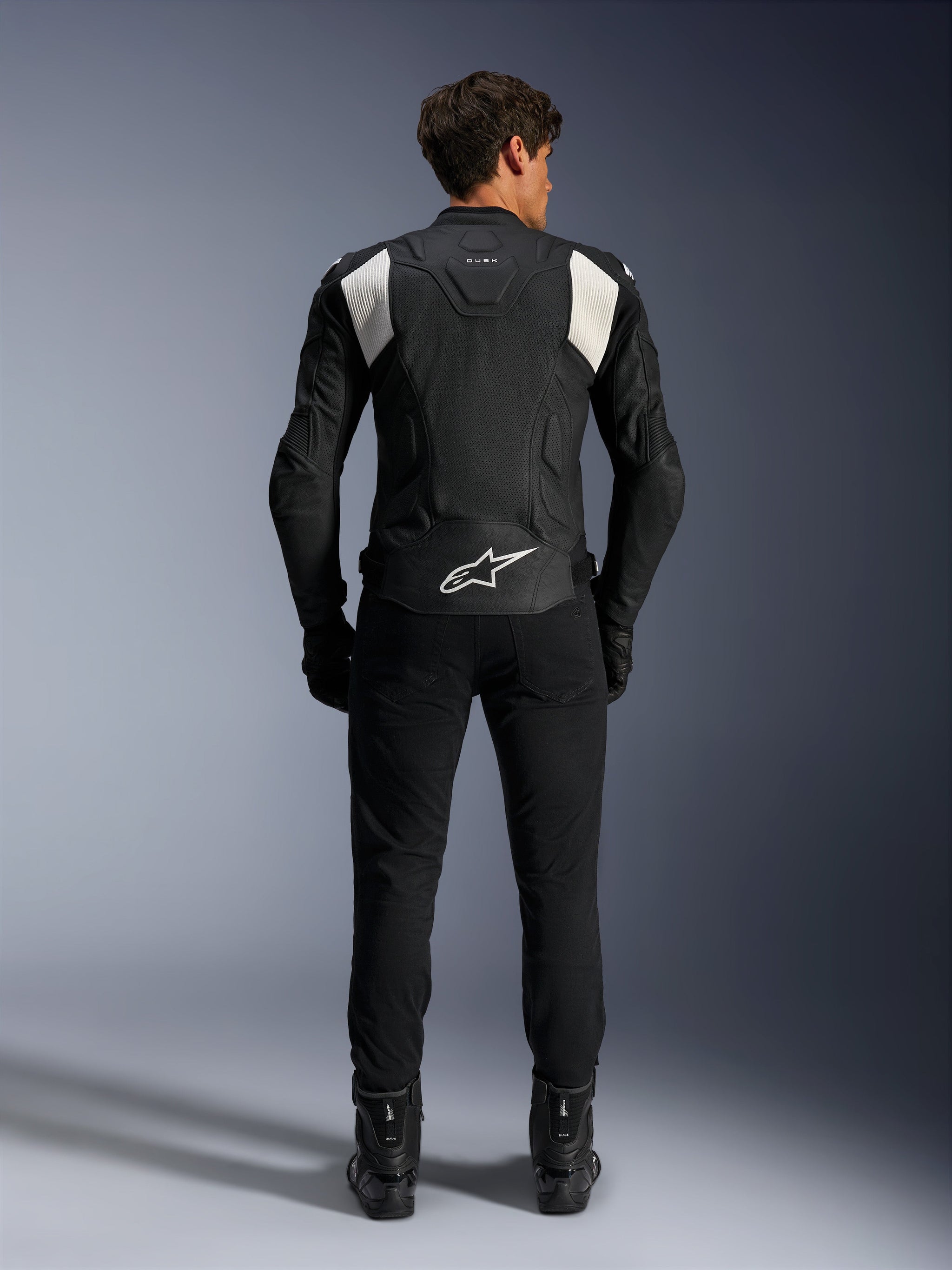 Alpinestars Dusk Airflow Leather Jacket, Black and White, rear view of a rider wearing black pants and boots, featuring perforated leather for ventilation, white accordion stretch panels on the shoulders, and white branding on the lower back