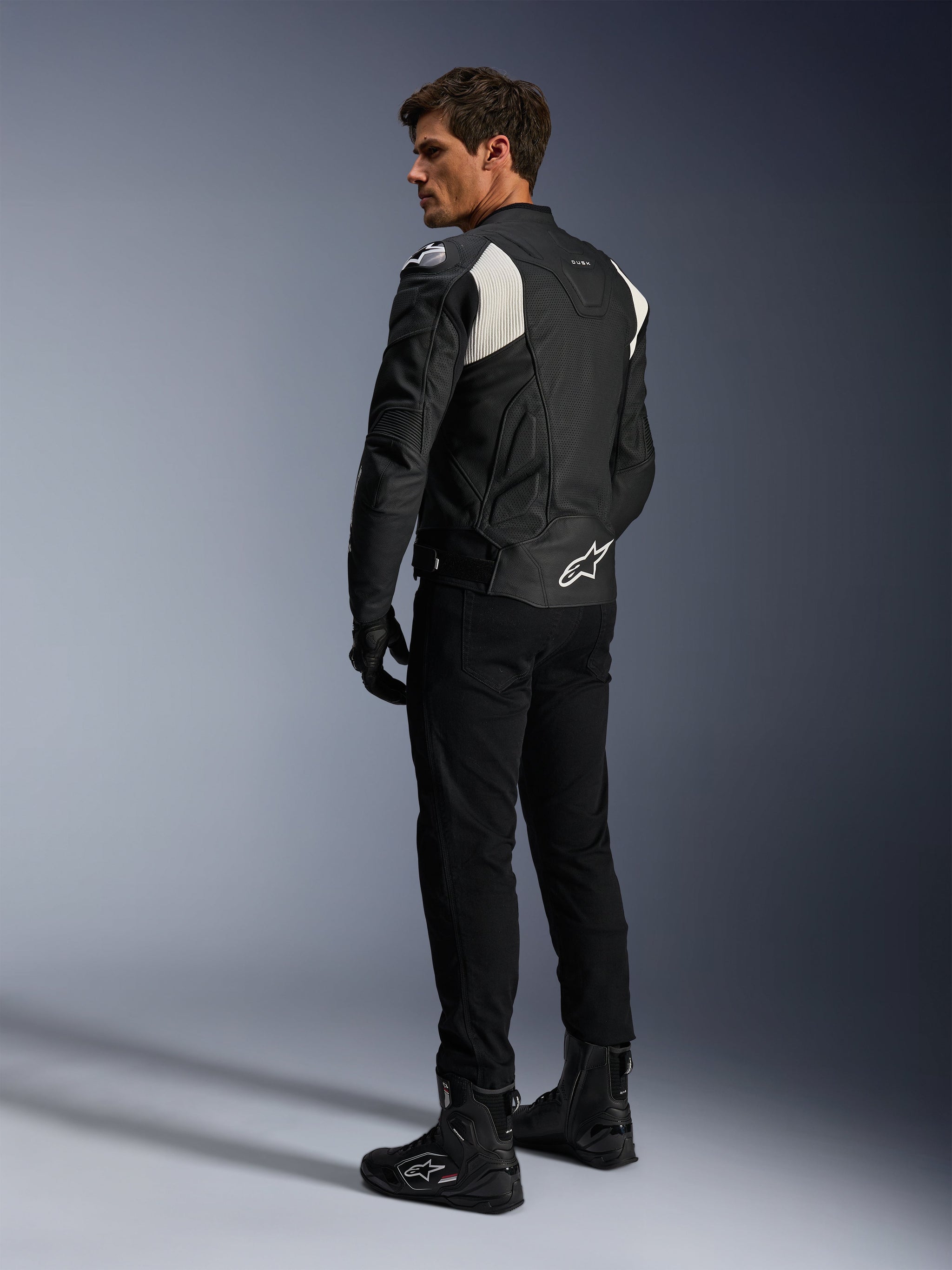 Alpinestars Dusk Airflow Leather Jacket, Black and White, rear view of model wearing the perforated leather jacket with matching black pants and boots, featuring white accordion stretch panels and bold logo on the lower back, sport motorcycle riding gear