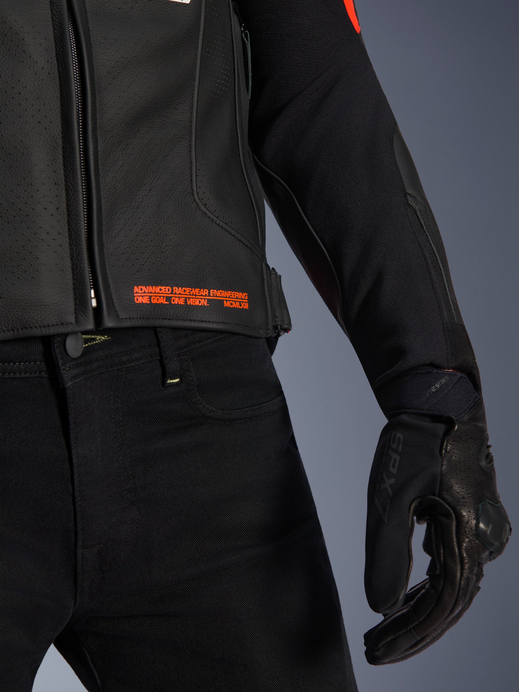 Alpinestars GP Plus R V4 Airflow Leather Jacket, close-up of waist and sleeve, black perforated leather with red fluo text branding reading 'Advanced Racewear Engineering', paired with black riding pants and SPX 7 motorcycle glove