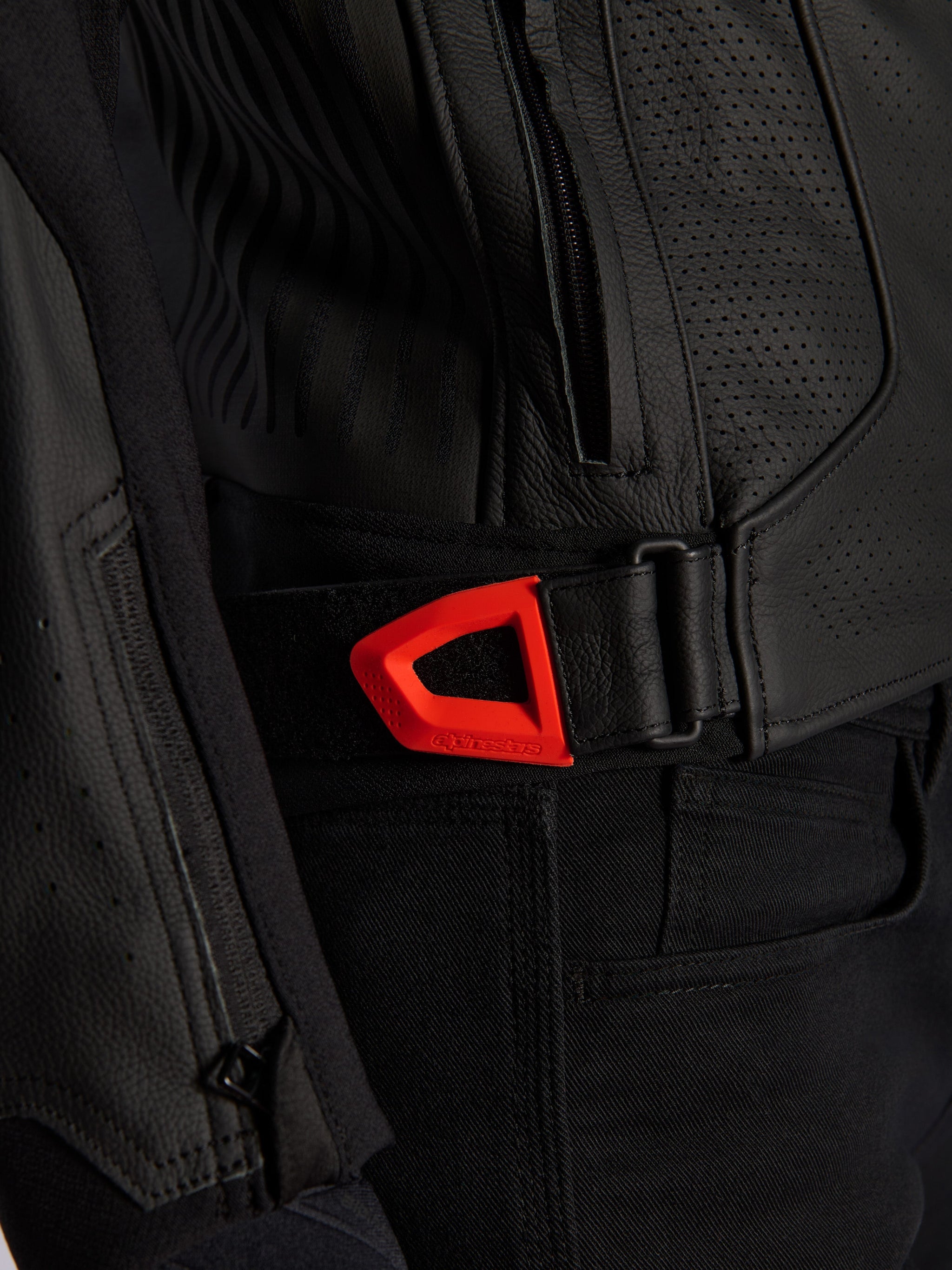 Alpinestars GP Plus R V4 Airflow Leather Jacket close-up, black perforated leather with a vibrant red fluo hook-and-loop waist adjustment strap, featuring a zippered side pocket and ergonomic textured panels.