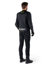 Rear view of a rider wearing the Alpinestars Faster V3 Airflow Leather Jacket, Black with White and Yellow Fluo accents, paired with black pants and boots, featuring perforated leather for ventilation and bold 'RACING AHEAD' branding on the lower back.