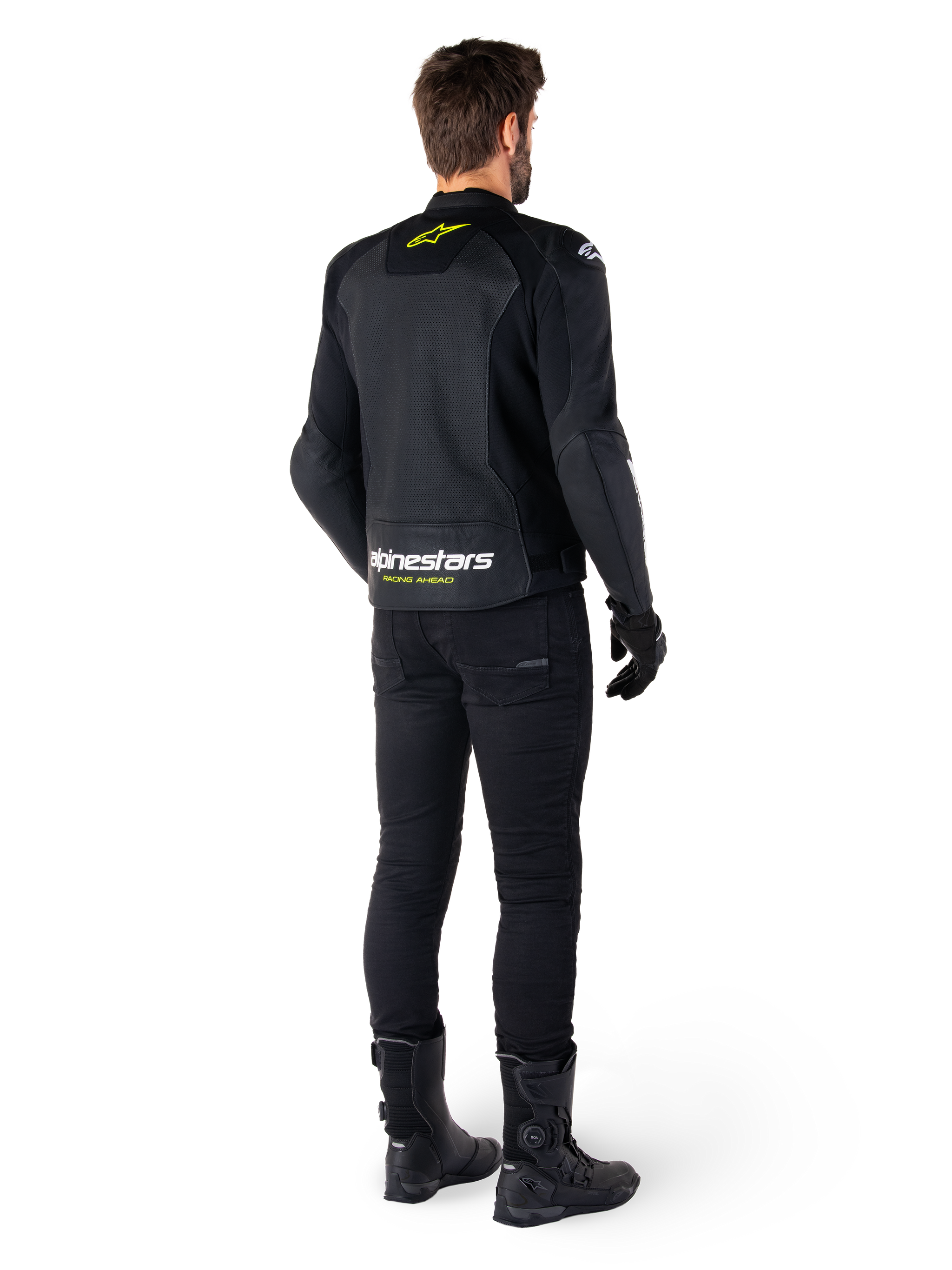 Rear view of a rider wearing the Alpinestars Faster V3 Airflow Leather Jacket, Black with White and Yellow Fluo accents, paired with black pants and boots, featuring perforated leather for ventilation and bold 'RACING AHEAD' branding on the lower back.