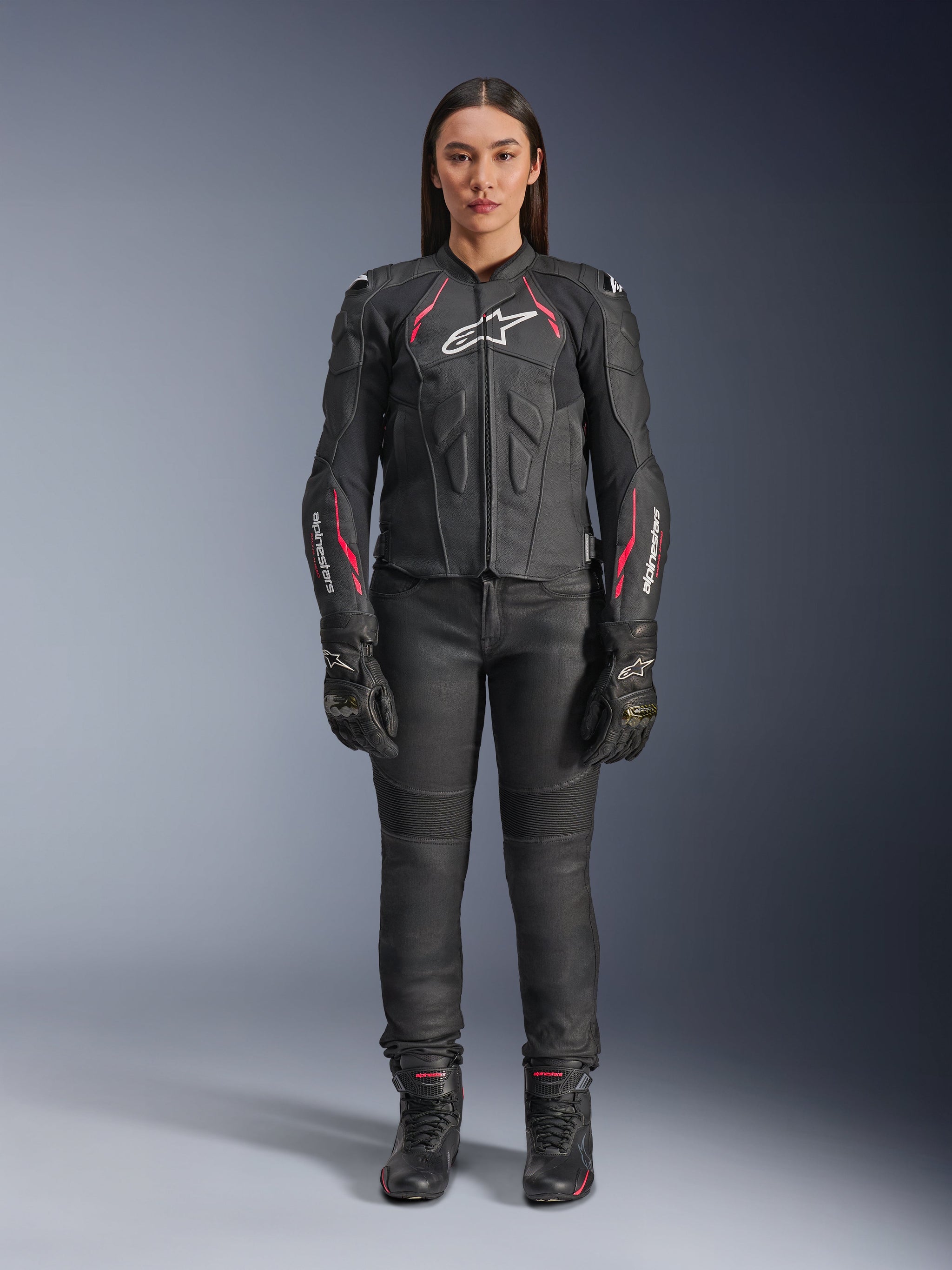 A woman wearing the Alpinestars Stella Dusk Leather Jacket in Black Diva Pink White, featuring a white chest logo and pink accents on the shoulders and sleeves, paired with black motorcycle pants, gloves, and boots.