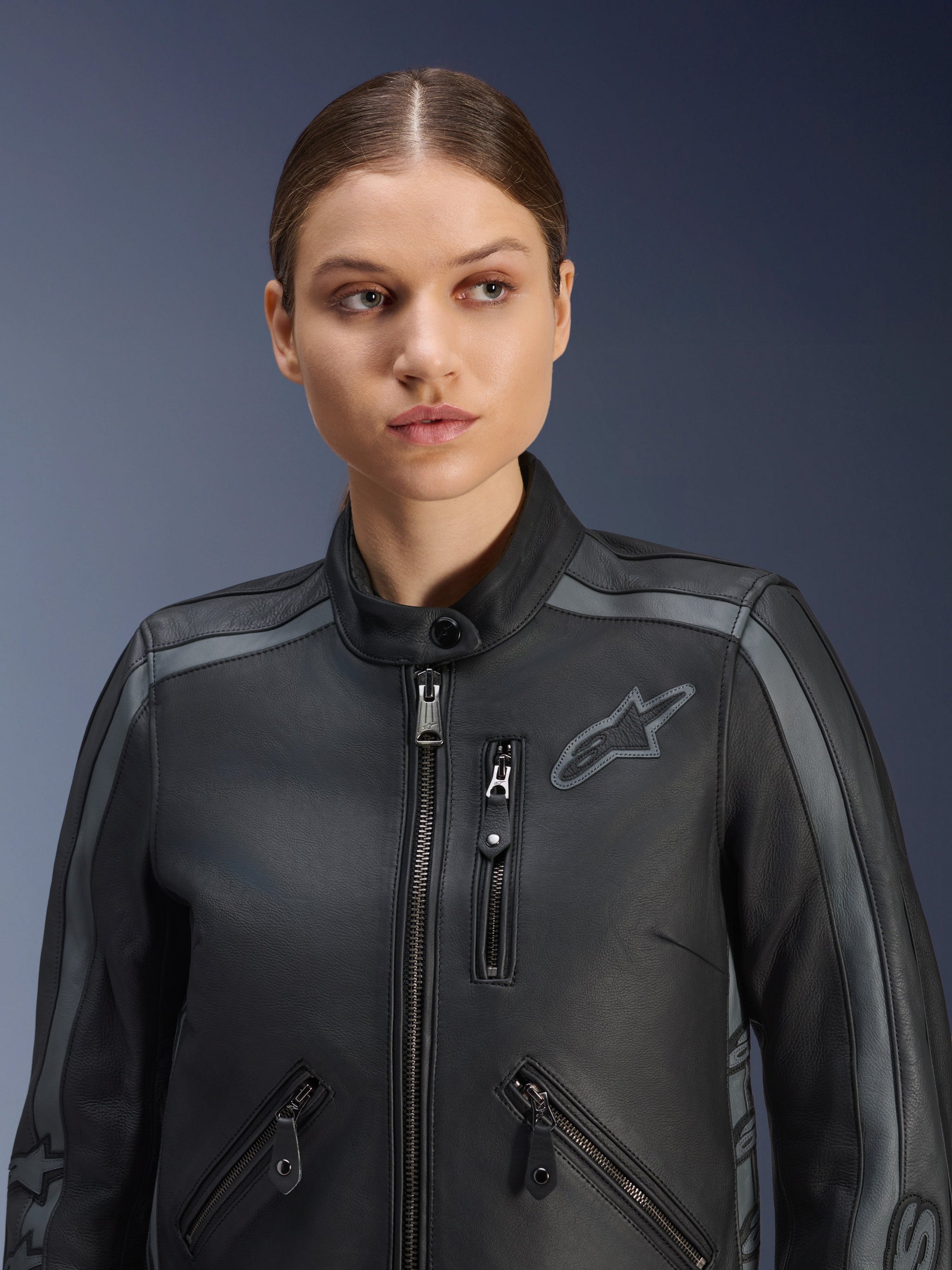 Female model wearing the Alpinestars Stella Dyno Leather Jacket in Black Tar Gray Dark Gray, premium bovine leather motorcycle jacket with a heritage design, featuring gray shoulder accents and multiple zippered pockets, tailored for a women's specific fit.