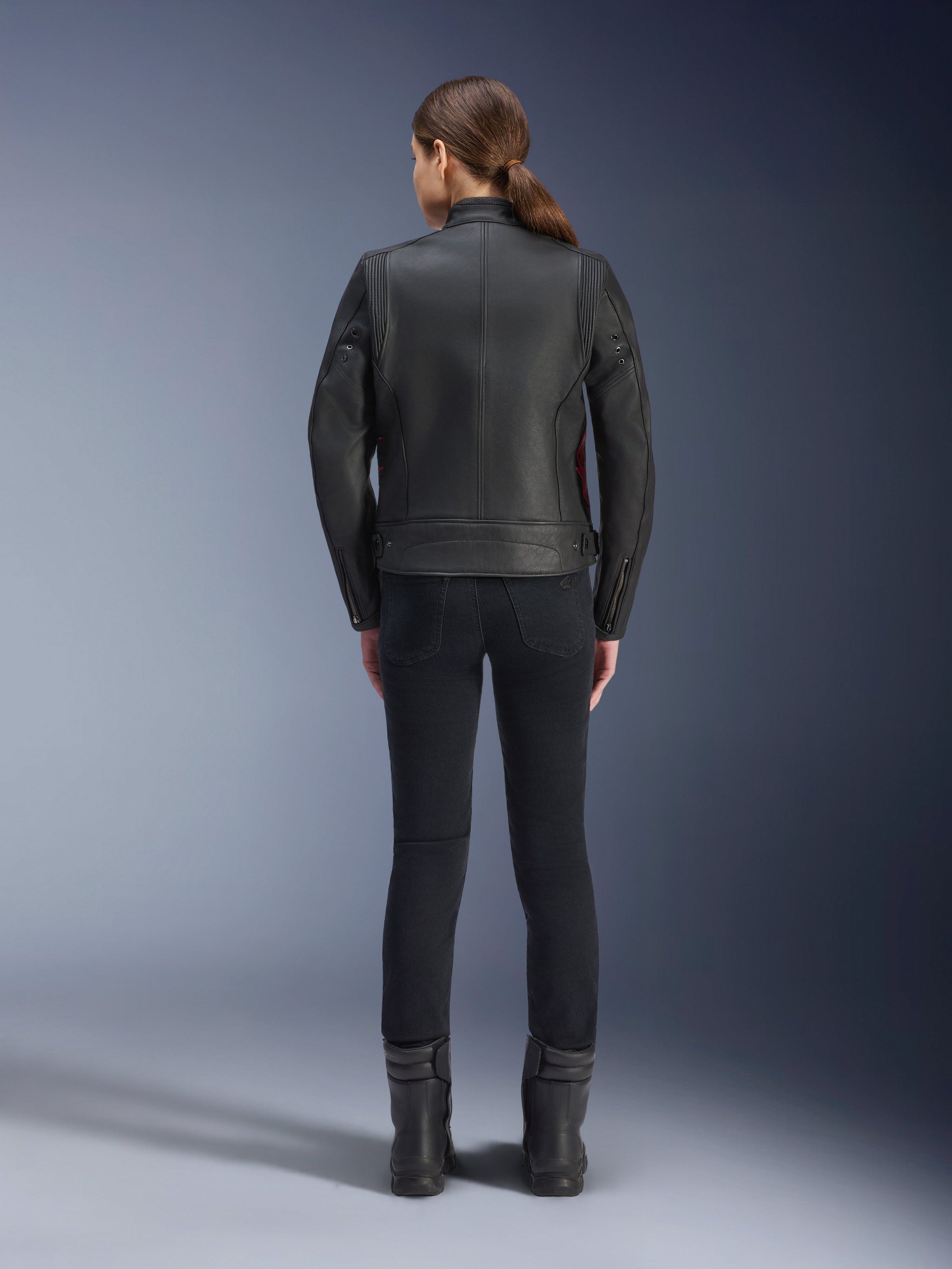 Rear view of a woman wearing the Alpinestars Stella Dyno Leather Jacket, Black with Haute Red accents, paired with slim-fit black pants and black boots, modern heritage leather motorcycle jacket design.