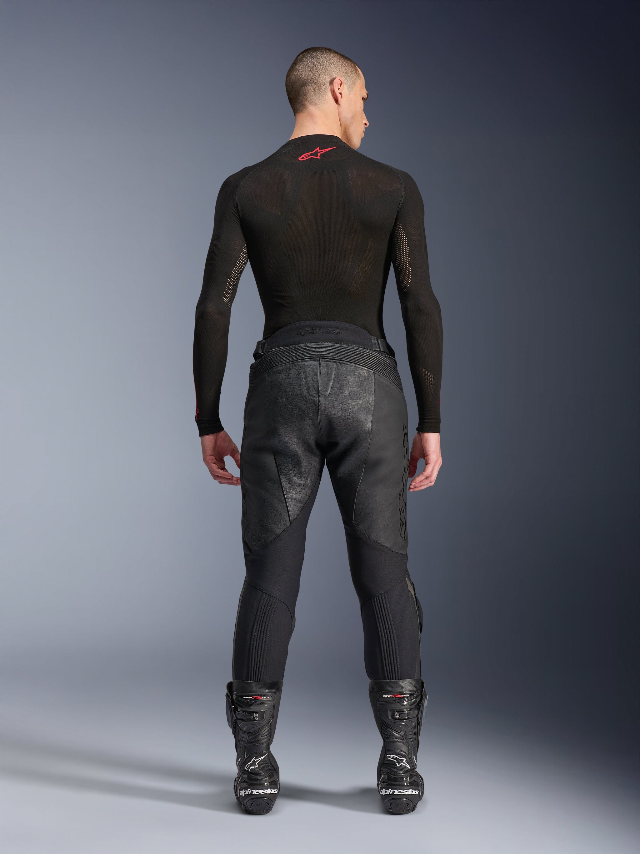 Alpinestars Missile V3 Leather Pants, Black, rear view on model wearing a black long-sleeve compression shirt and racing boots, premium leather construction with high-waist overlap and stretch zones, professional motorcycle racing gear