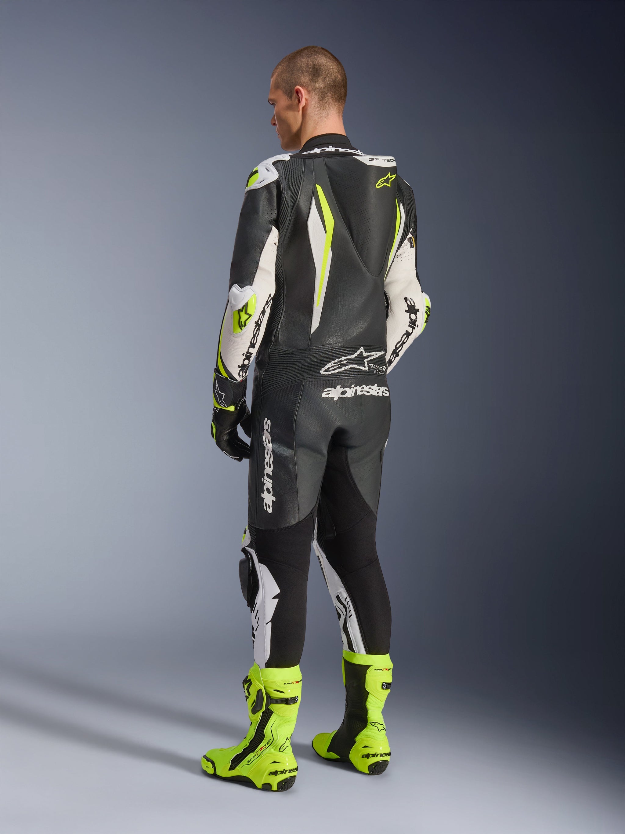 GP Tech V5 One Piece Leather Suit, Black White and Yellow Fluo, back view of rider wearing full racing suit with matching fluorescent yellow boots, featuring an aerodynamic speed hump and bold Alpinestars logos, professional motorcycle gear
