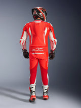 Rear view of a rider wearing the Alpinestars GP Tech V5 One Piece Leather Suit in Red Fluo, Bright Red, and White. High-performance leather suit with an aerodynamic speed hump, white accents, and Alpinestars branding, paired with a matching helmet, black gloves, and white and red racing boots.