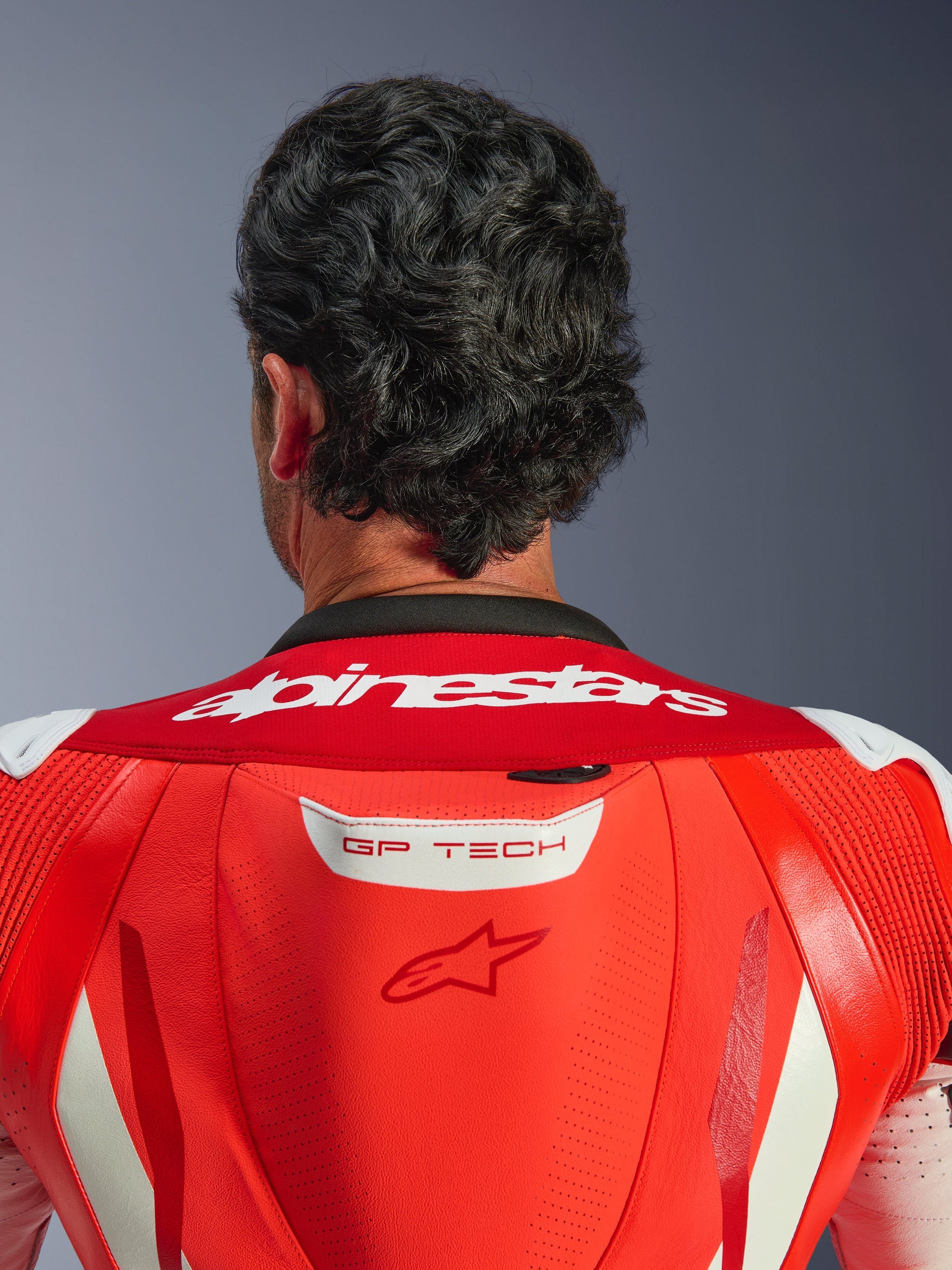 Close-up of the upper back and neck of the GP Tech V5 One Piece Leather Suit, Red Fluo and Bright Red, featuring white Alpinestars wordmark on the collar and GP TECH logo on the aerodynamic hump, worn by a rider.