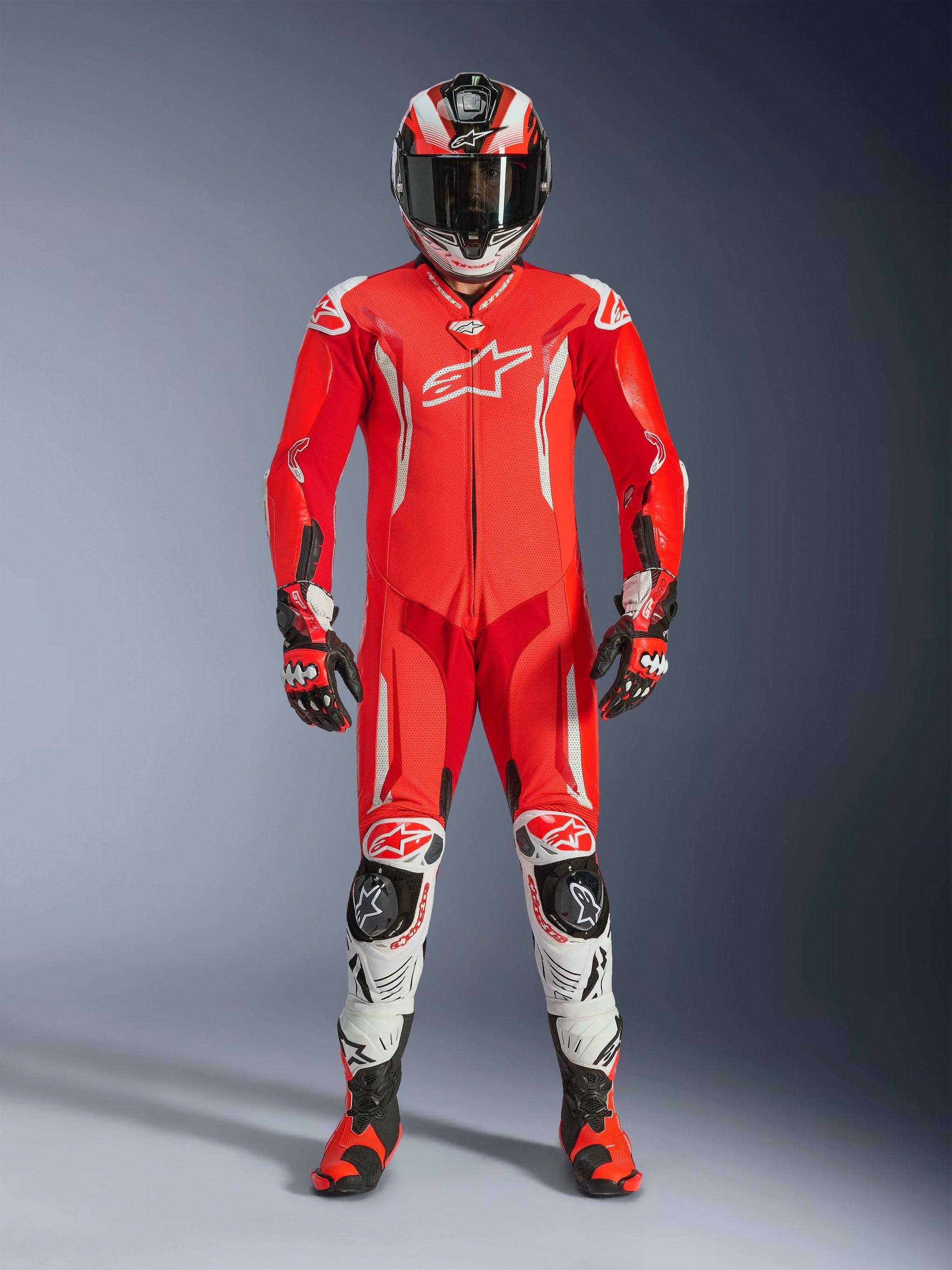 Rider wearing the Alpinestars GP Tech V5 One Piece Leather Suit in Red Fluo, Bright Red, and White, complete with a matching racing helmet, gloves, and boots. Professional track suit featuring an aerodynamic silhouette, white Alpinestars logos, and integrated protective sliders.