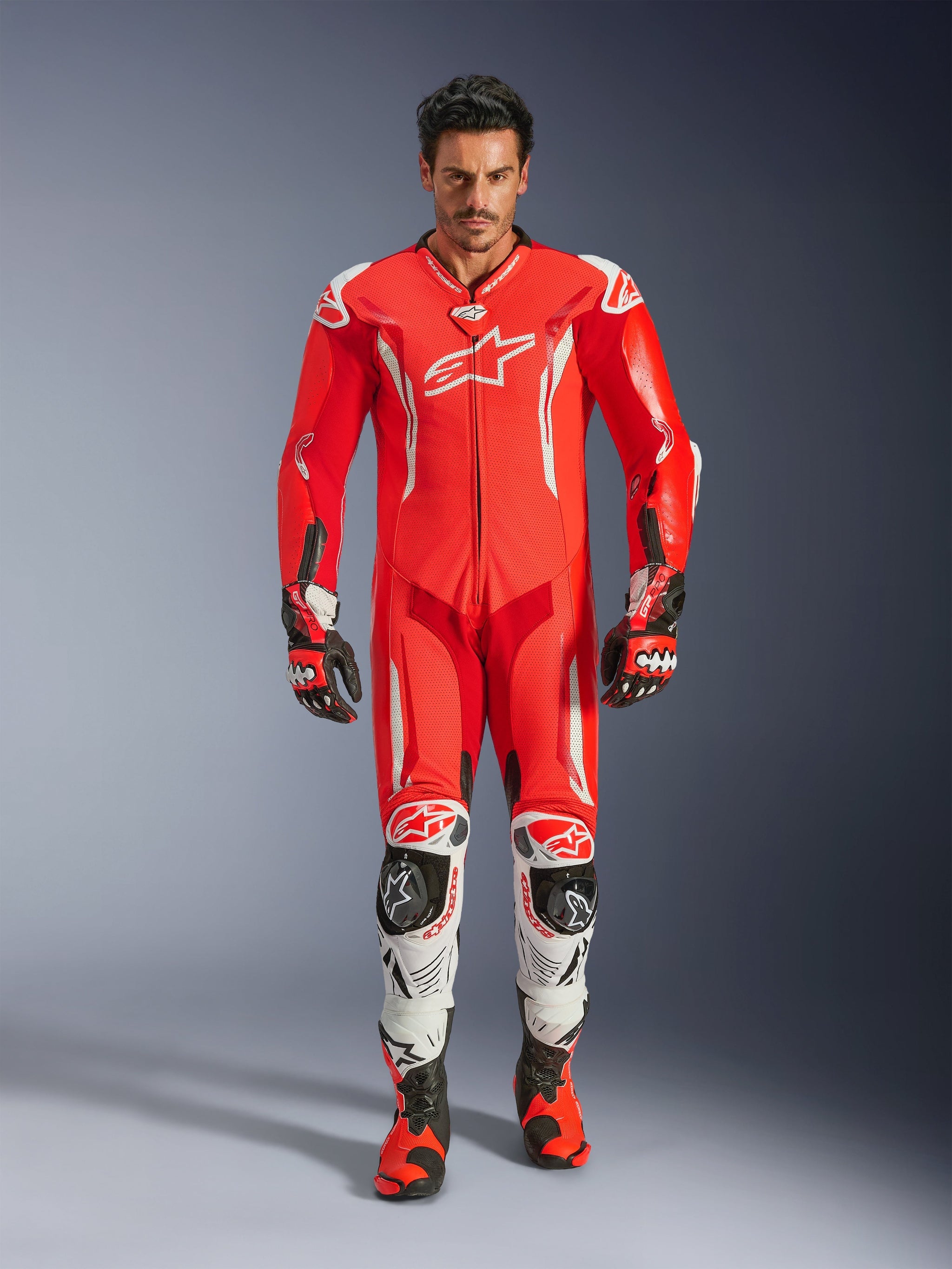 Alpinestars GP Tech V5 One Piece Leather Suit, professional racing suit in Red Fluo and Bright Red with white accents, worn by a rider with matching boots and gloves, featuring an aerodynamic speed hump, premium leather construction, and advanced protective armor for track performance.