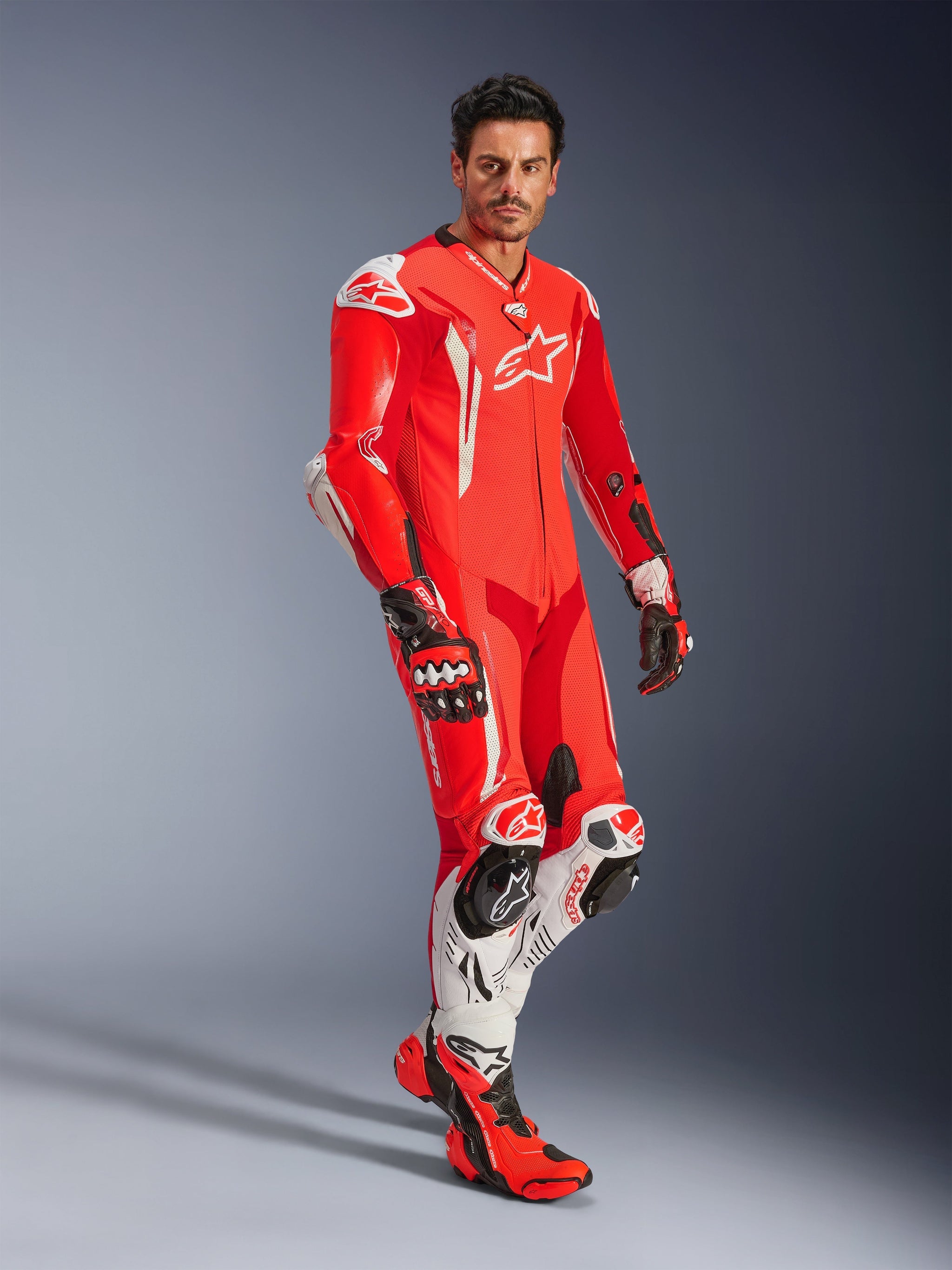 Man wearing Alpinestars GP Tech V5 One Piece Leather Suit, Red Fluo Bright Red and White, coordinated with racing gloves and boots, professional motorcycle racing gear featuring aerodynamic humps and integrated protection.