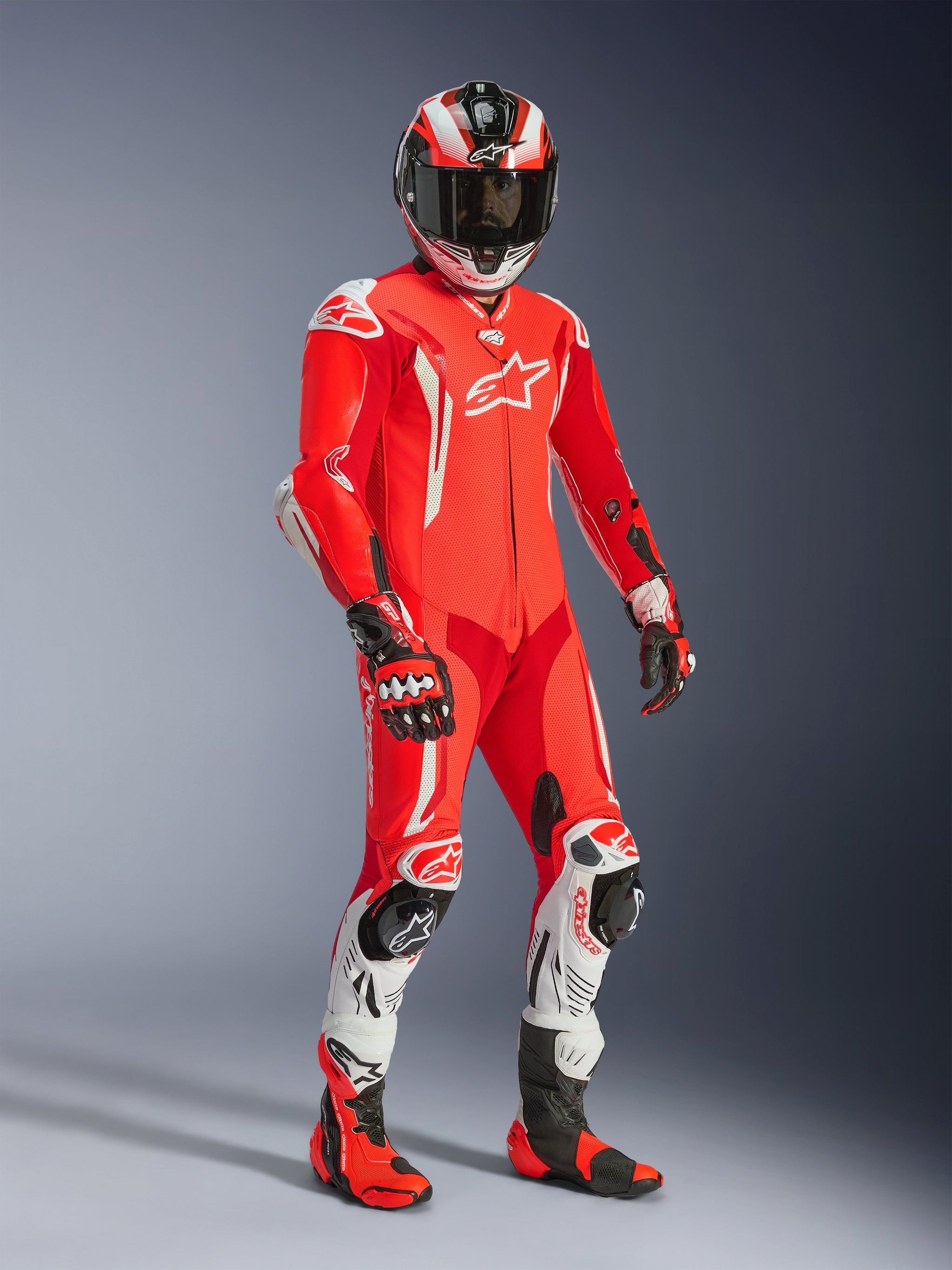 Motorcycle rider wearing the Alpinestars GP Tech V5 One Piece Leather Suit, Red Fluo and Bright Red with White accents, featuring matching helmet, gloves, and racing boots, professional track gear with perforated leather and protective sliders