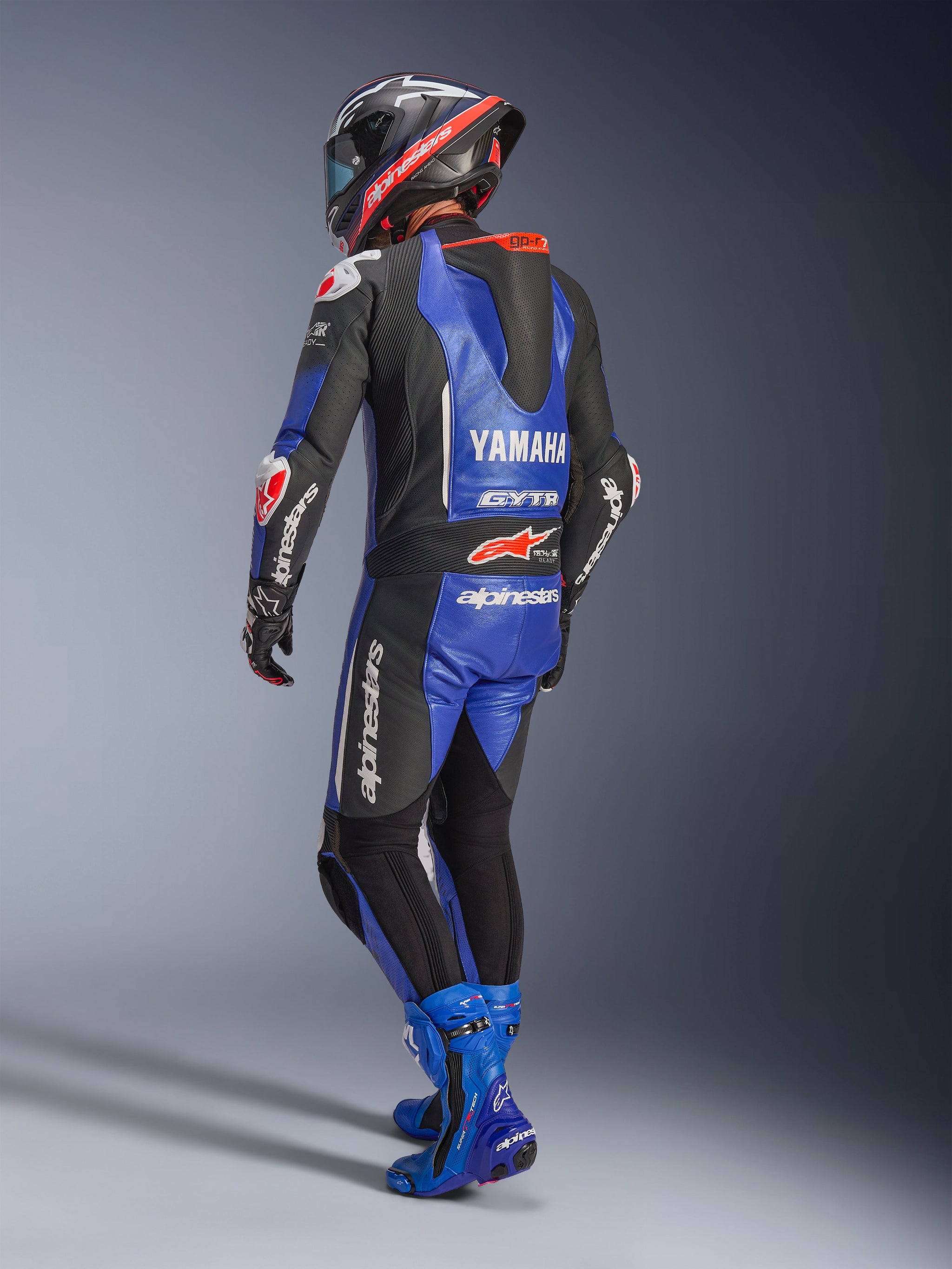 Alpinestars Yamaha GP-R7 One Piece Leather Suit, Metallic Blue Black and White, Rear view showing aerodynamic race hump and Yamaha branding, worn with matching blue racing boots and helmet, professional performance leather suit for track racing