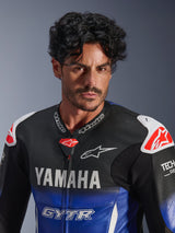 Close-up portrait of a man wearing the Alpinestars Yamaha GP-R7 One Piece Leather Suit, Metallic Blue and Black with white YAMAHA and GYTR branding, perforated leather, white shoulder sliders with red star logos, Tech-Air Ready, professional motorcycle racing gear.