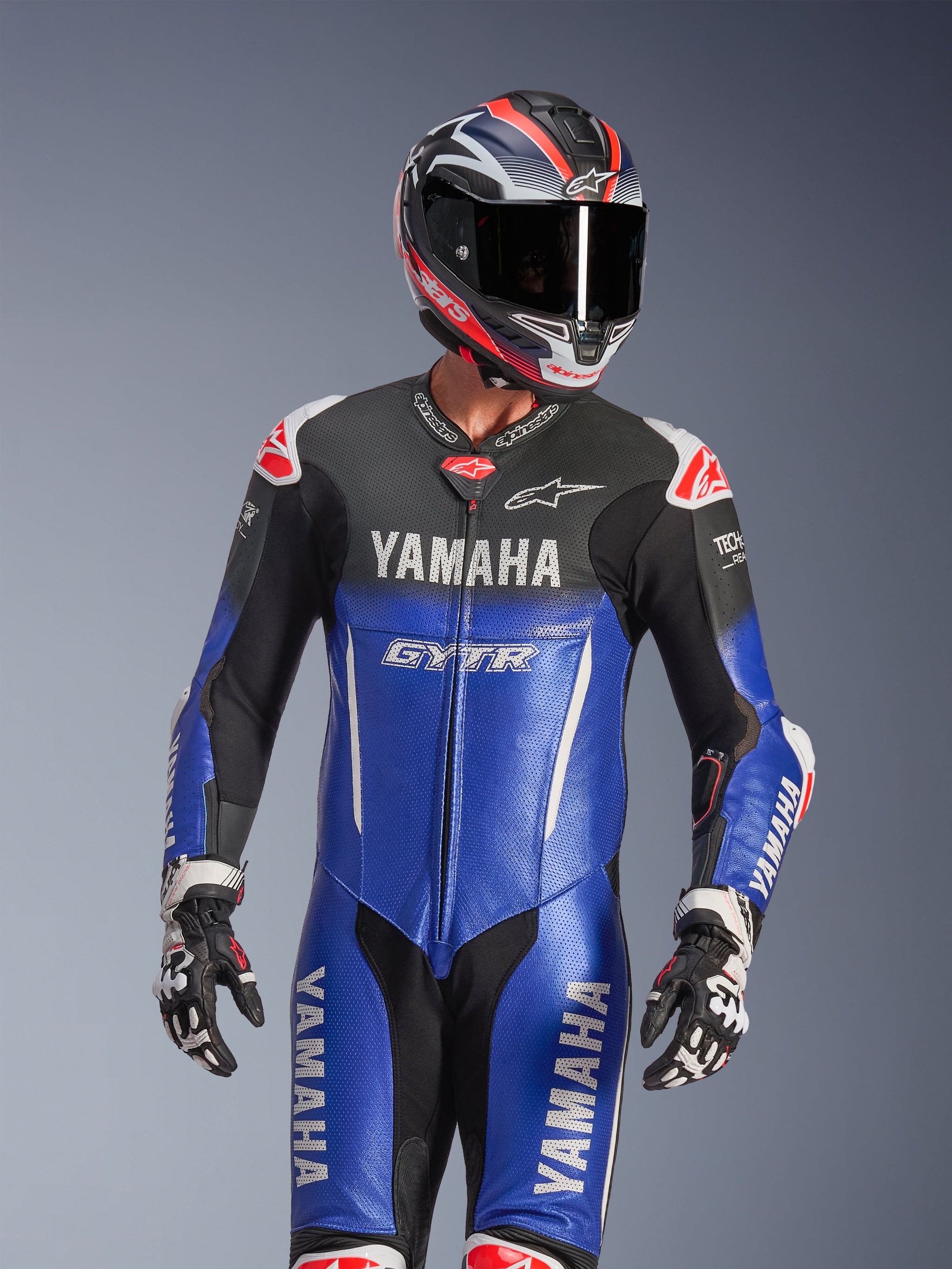 Yamaha GP-R7 One Piece Leather Suit, Metallic Blue and Black with White and Red accents, worn with a matching helmet and racing gloves, featuring YAMAHA and GYTR branding, perforated leather panels, and Alpinestars logos, professional motorcycle racing apparel.