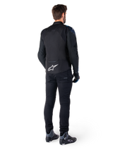 Rear view of a man wearing the Alpinestars T-Jaws V4 Waterproof Jacket in Black, Dark Blue, and Blithe Blue, featuring a prominent white Astar logo on the lower back, paired with black pants and motorcycle riding shoes.