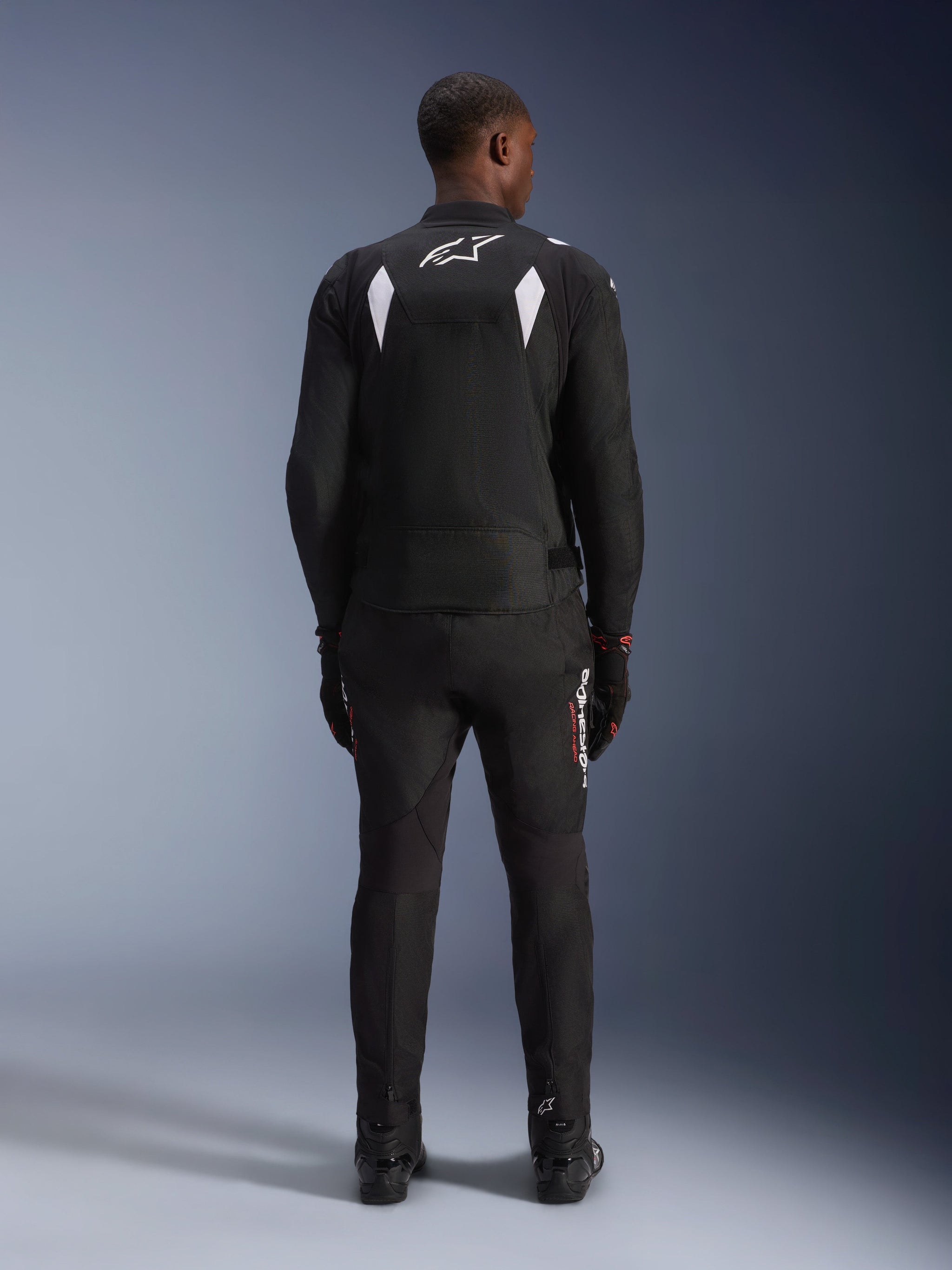 Alpinestars T-SP 1 V2 Waterproof Jacket, Black and White, rear view of a rider wearing the jacket with matching black motorcycle pants and boots, featuring a white A-star logo on the upper back and reflective shoulder panels, sport textile riding gear
