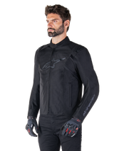 Model wearing Alpinestars T-SPS V2 Waterproof Jacket, Black, paired with black pants and grey motorcycle gloves with red accents, featuring a subtle black chest logo and sleeve branding, textile sport-cut jacket for road riding.