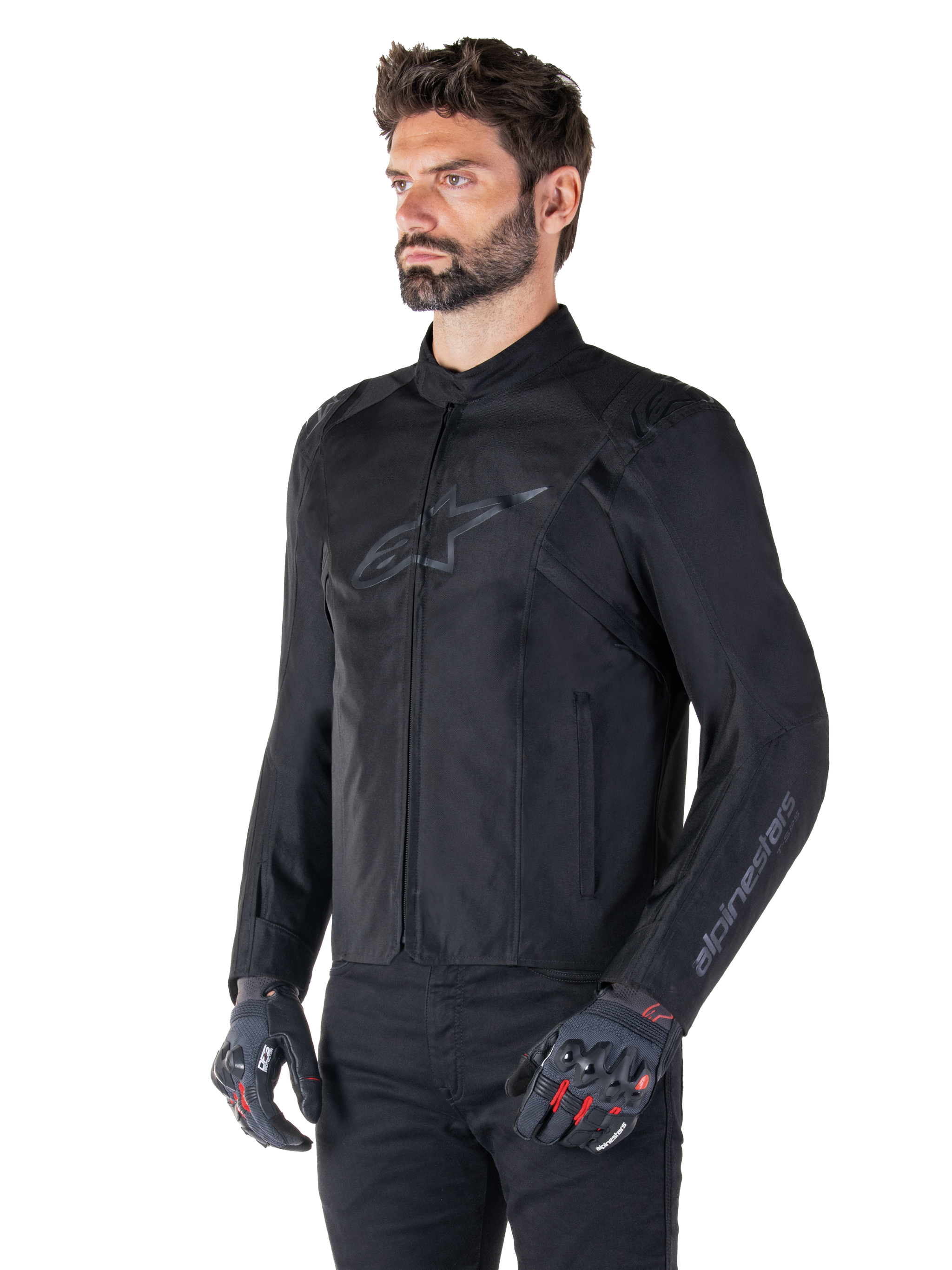 Model wearing Alpinestars T-SPS V2 Waterproof Jacket, Black, paired with black pants and grey motorcycle gloves with red accents, featuring a subtle black chest logo and sleeve branding, textile sport-cut jacket for road riding.