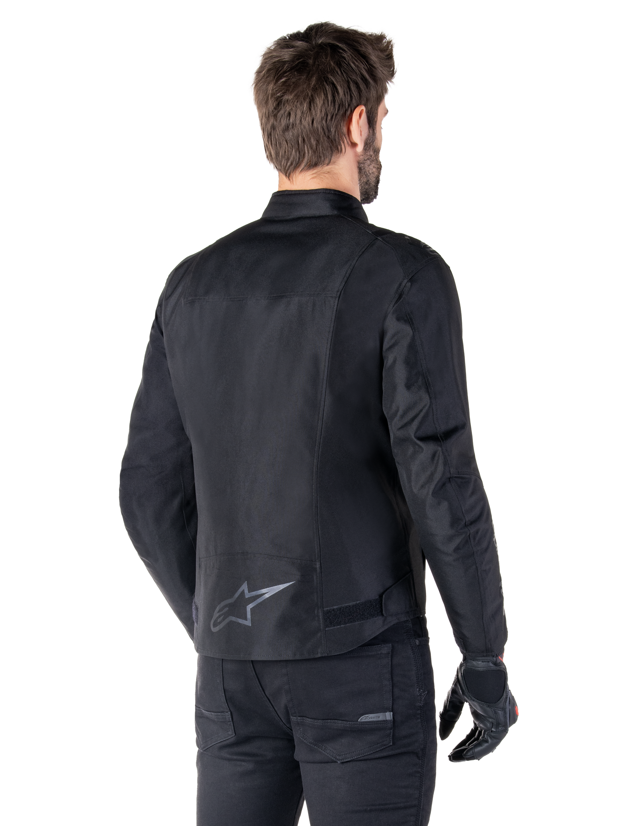 Alpinestars T-SPS V2 Waterproof Jacket, Black, rear view on a male model, featuring a reflective star logo on the lower back, extended back profile, and waist adjustment straps, paired with black riding pants and gloves