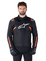 Alpinestars T-SPS V2 Waterproof Jacket, Black White and Red Fluo, worn by a male model with black riding gloves, featuring a large white Alpinestars logo on the chest and vibrant orange accents on the shoulders and sleeves.