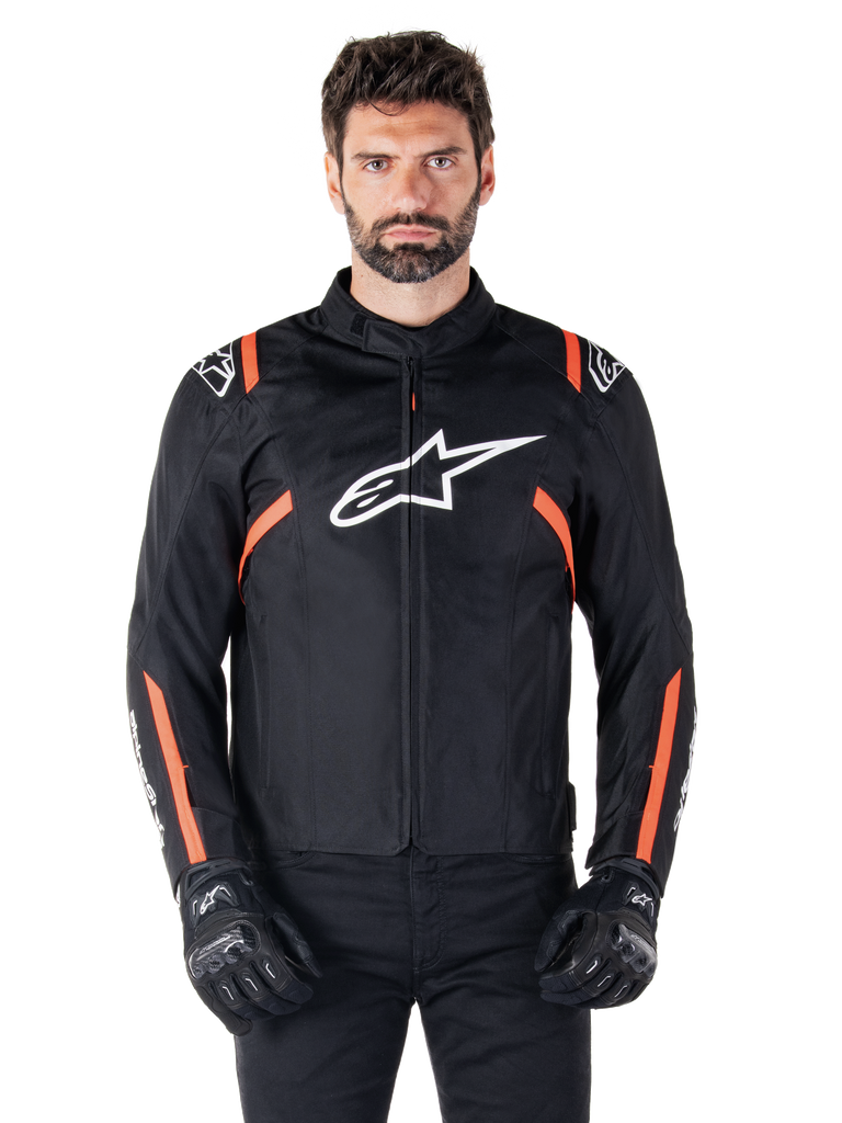 Alpinestars T-SPS V2 Waterproof Jacket, Black White and Red Fluo, worn by a male model with black riding gloves, featuring a large white Alpinestars logo on the chest and vibrant orange accents on the shoulders and sleeves.