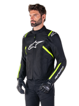 Male model wearing the Alpinestars T-SPS V2 Waterproof Jacket, Black with White and Yellow Fluo accents, featuring a white Astars logo on the chest and fluorescent yellow stripes on the shoulders and sleeves, paired with black pants and riding gloves, sport-cut textile motorcycle jacket.