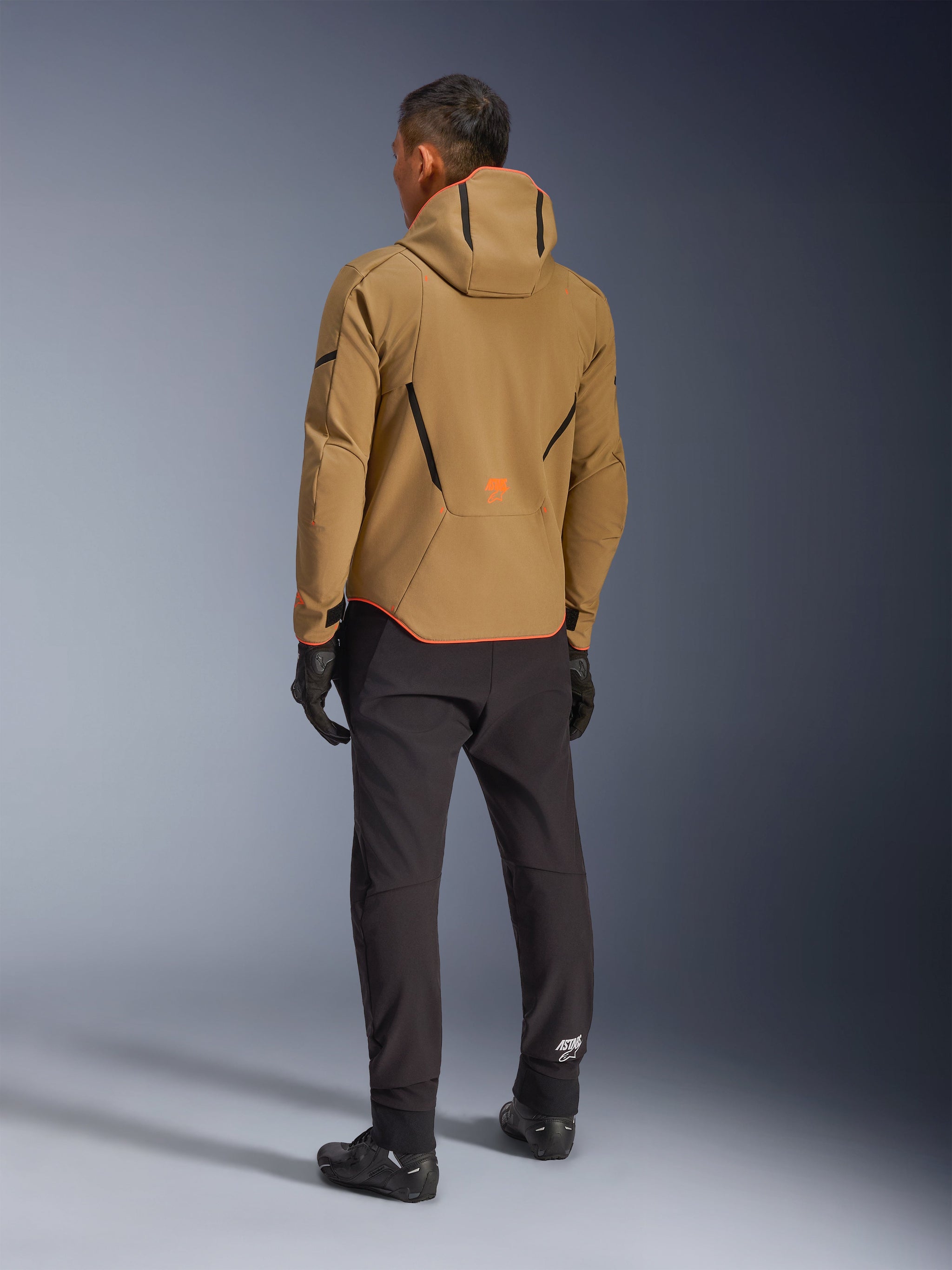 Alpinestars Aeroshell Waterproof Jacket, Dark Khaki Black Orange Fluo, back view of a rider wearing the hooded softshell jacket, paired with black pants and gloves, featuring black geometric accents and an orange logo on the lower back.