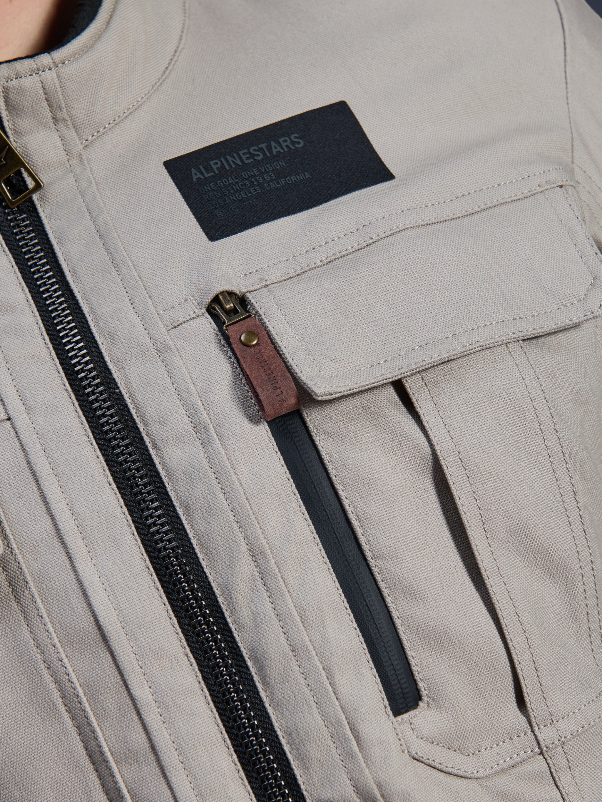 Alpinestars Borrego Drystar® Jacket, Warm Gray, close-up detail of chest area featuring a black Alpinestars branding patch, chest pocket with flap, and a vertical zipper with a brown leather pull tab, durable gray textile canvas construction.