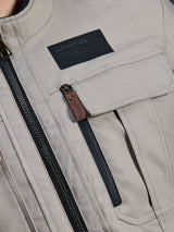 Alpinestars Borrego Drystar® Jacket, Warm Gray, close-up detail of chest area featuring a black Alpinestars branding patch, chest pocket with flap, and a vertical zipper with a brown leather pull tab, durable gray textile canvas construction.