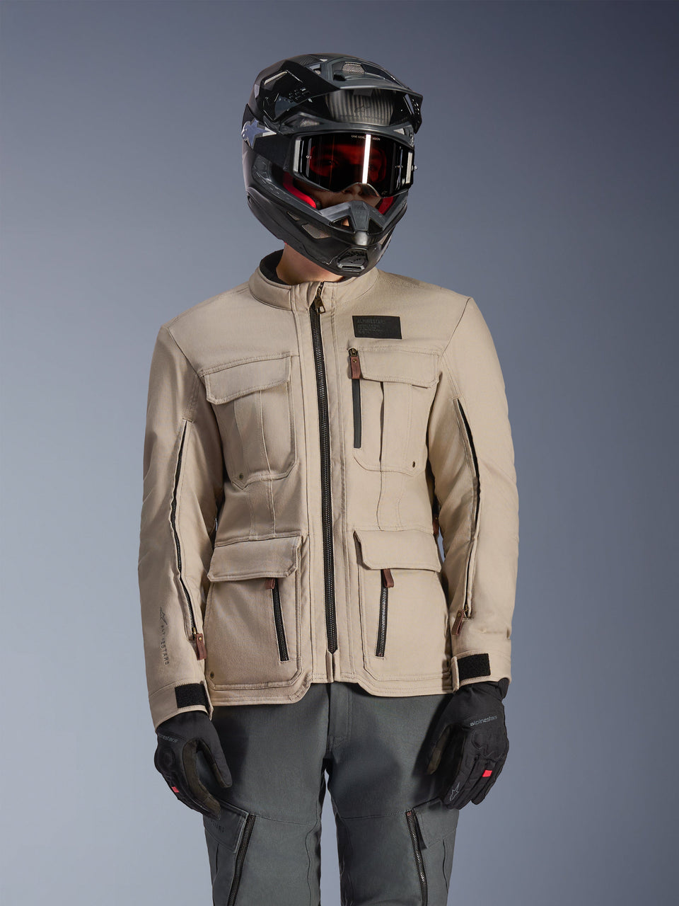 Alpinestars Borrego Drystar® Jacket, Adventure Motorcycle Jacket, Warm Gray, featuring multiple front cargo pockets and black zippers, worn with a black helmet, goggles, and dark gray riding pants, rugged textile design for touring