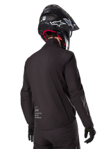 Alpinestars AMT-8 Stretch Drystar® XF Liner, Black Waterproof Jacket Liner, rear view on a rider wearing a black helmet and gloves, featuring Alpinestars Motion Technology branding on the lower back, minimalist and functional design for adventure riding.