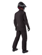 Rear view of a rider wearing the Alpinestars AMT-8 Stretch Drystar® XF Jacket liner, Black technical motorcycle gear, worn with matching pants and black helmet, featuring "AMT Alpinestars Motion Technology" branding on the back, sleek minimalist design for adventure touring