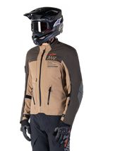 Alpinestars AMT-8 Stretch Drystar® XF Jacket, Adventure Motorcycle Jacket, Dark Brown and Dark Khaki, worn with a black off-road helmet and goggles, featuring Alpinestars Motion Technology branding and zippered ventilation ports.